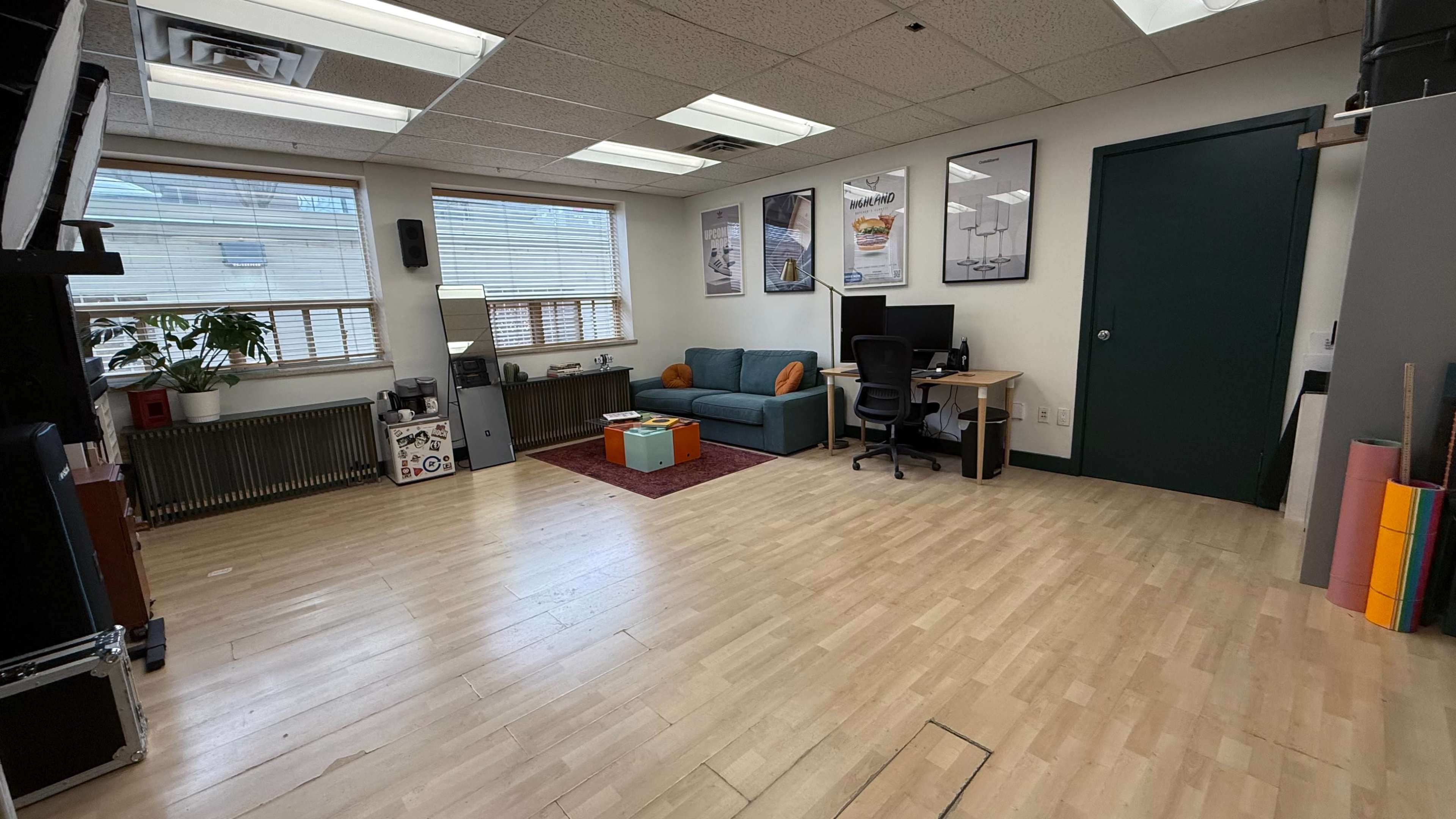 The image shows a spacious office or studio area with a teal couch, a computer desk, posters on the walls, and wooden flooring.