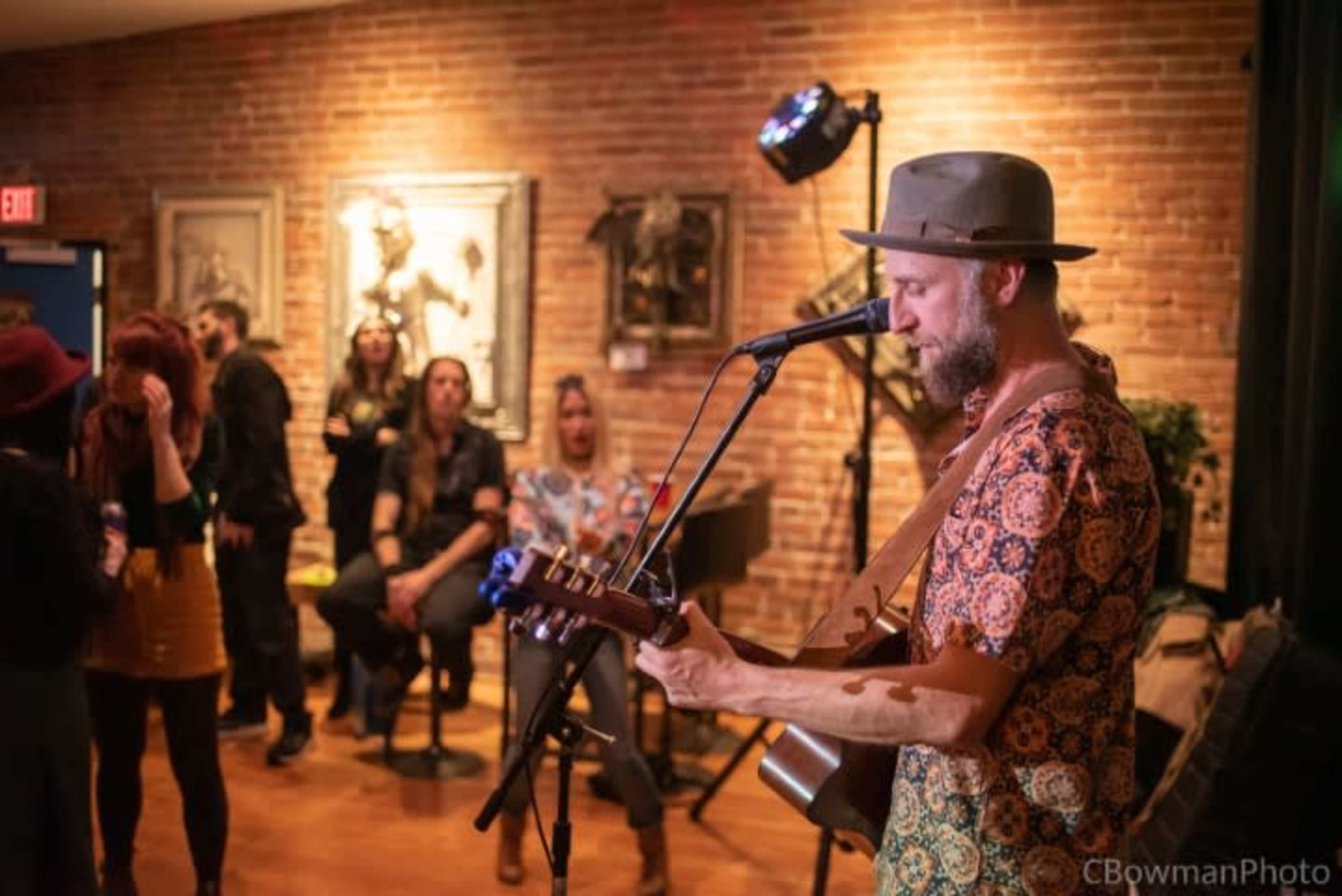 A musician performs with an acoustic guitar in a lively venue filled with spectators, while brick walls and soft lighting create an inviting atmosphere.