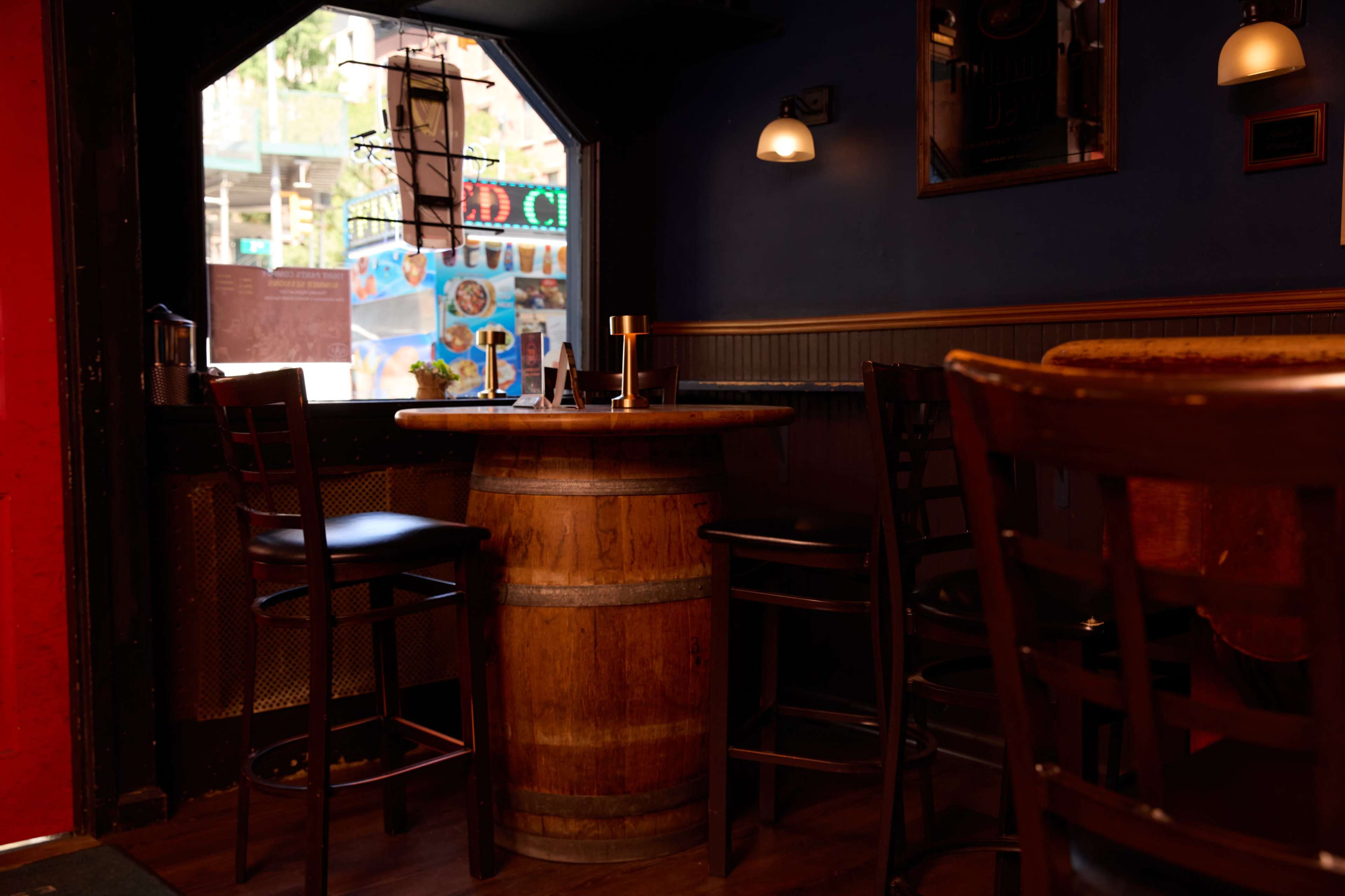 The image shows a cozy bar area with wooden furniture, including a barrel table and several high-backed chairs, next to a window overlooking a street scene.