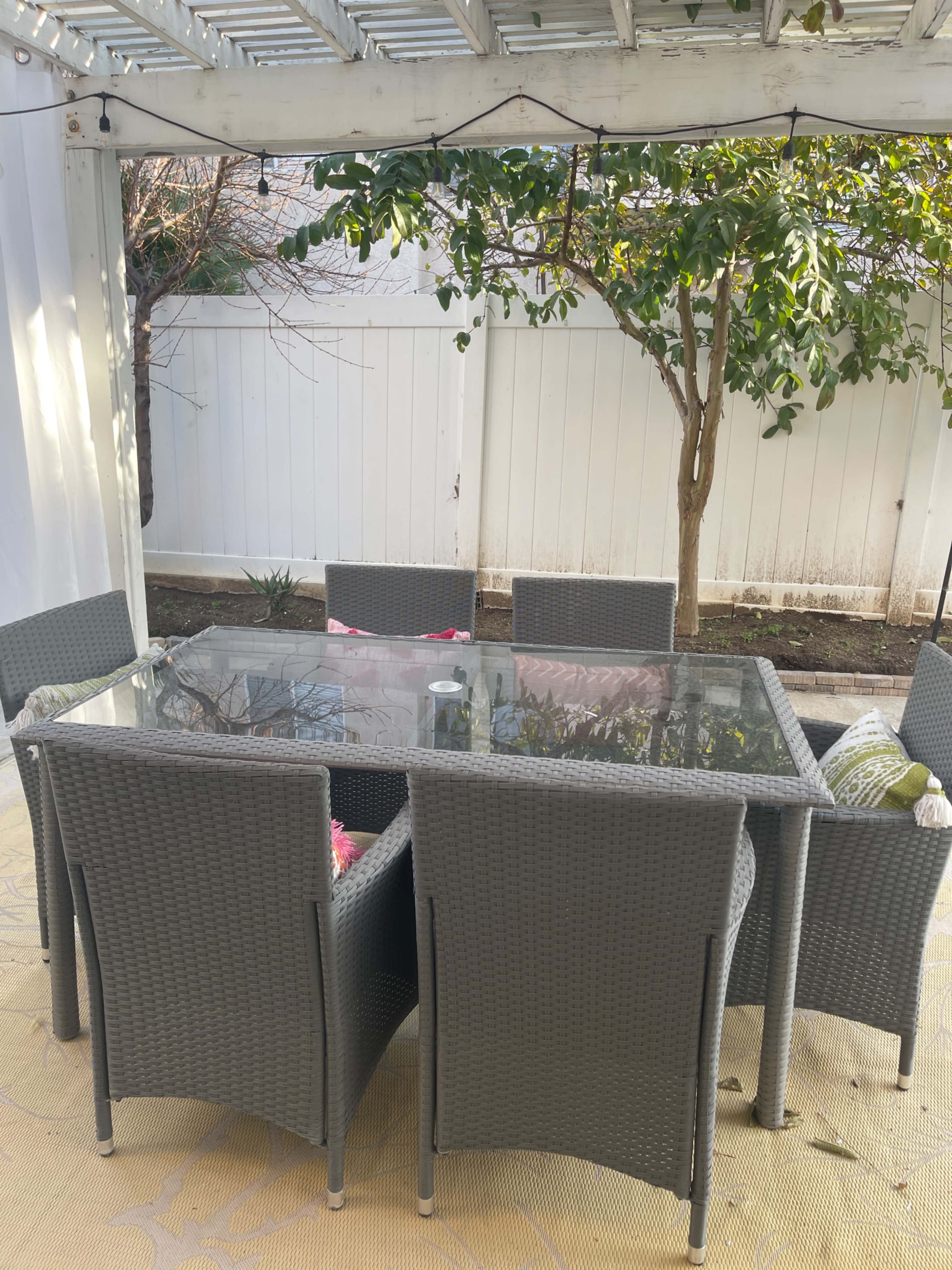 A rectangular glass table is surrounded by six wicker chairs on a patio under a covered structure, with a tree and fence in the background.