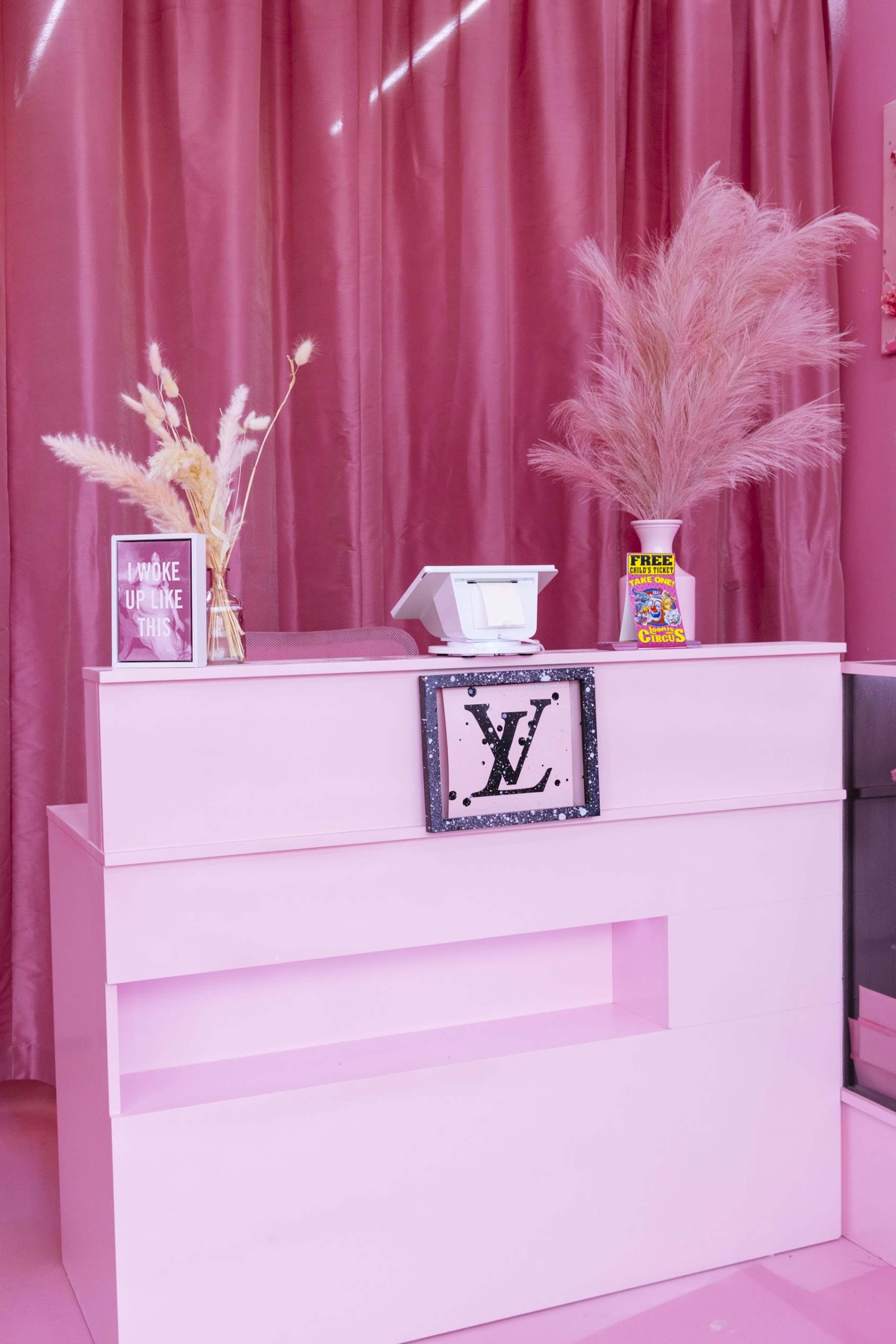 The image shows a pink-themed retail checkout area featuring a cash register, decorative pampas grass, and colorful merchandise.