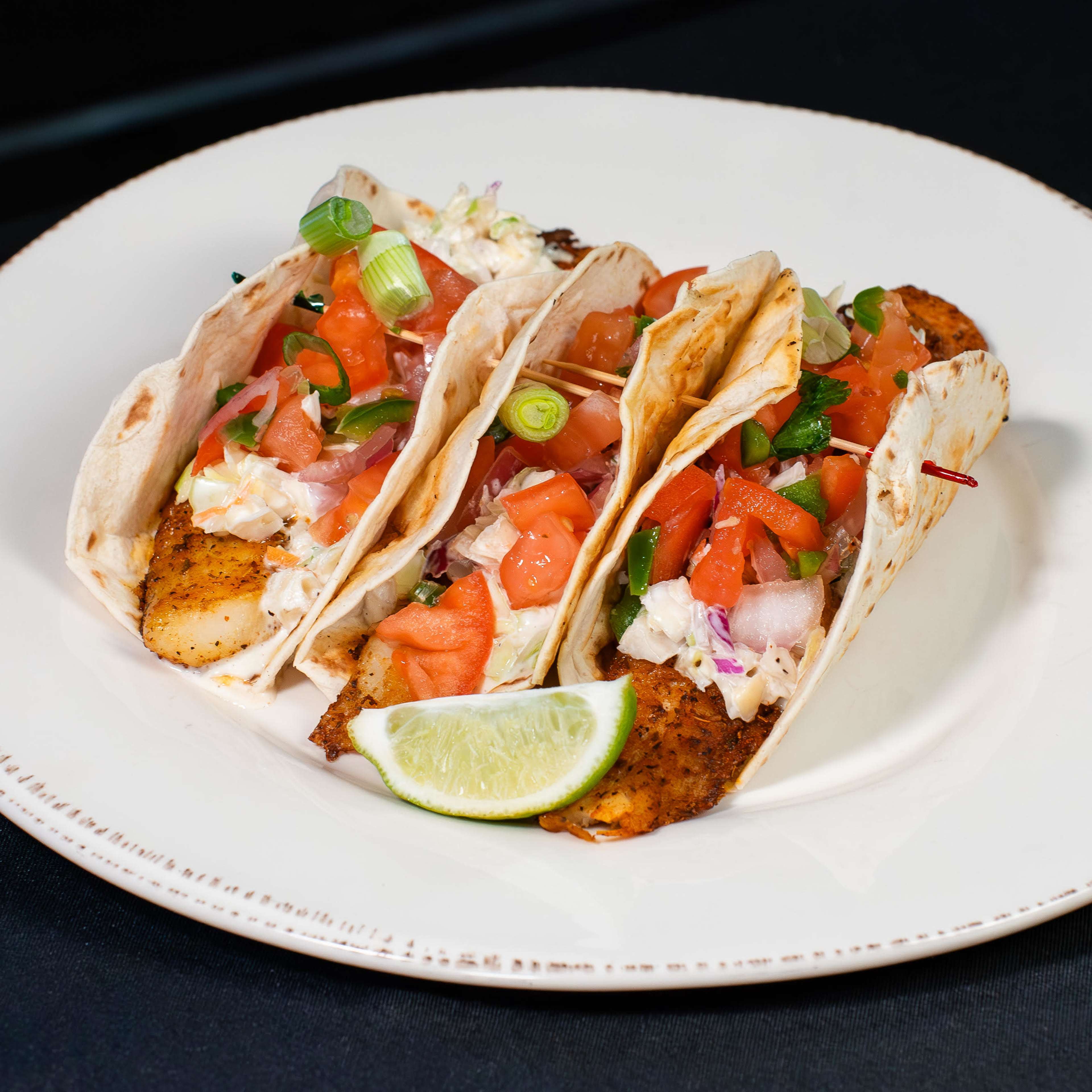 Three fish tacos are arranged on a white plate, filled with chopped tomatoes, green onions, and shredded cabbage, accompanied by a lime wedge.