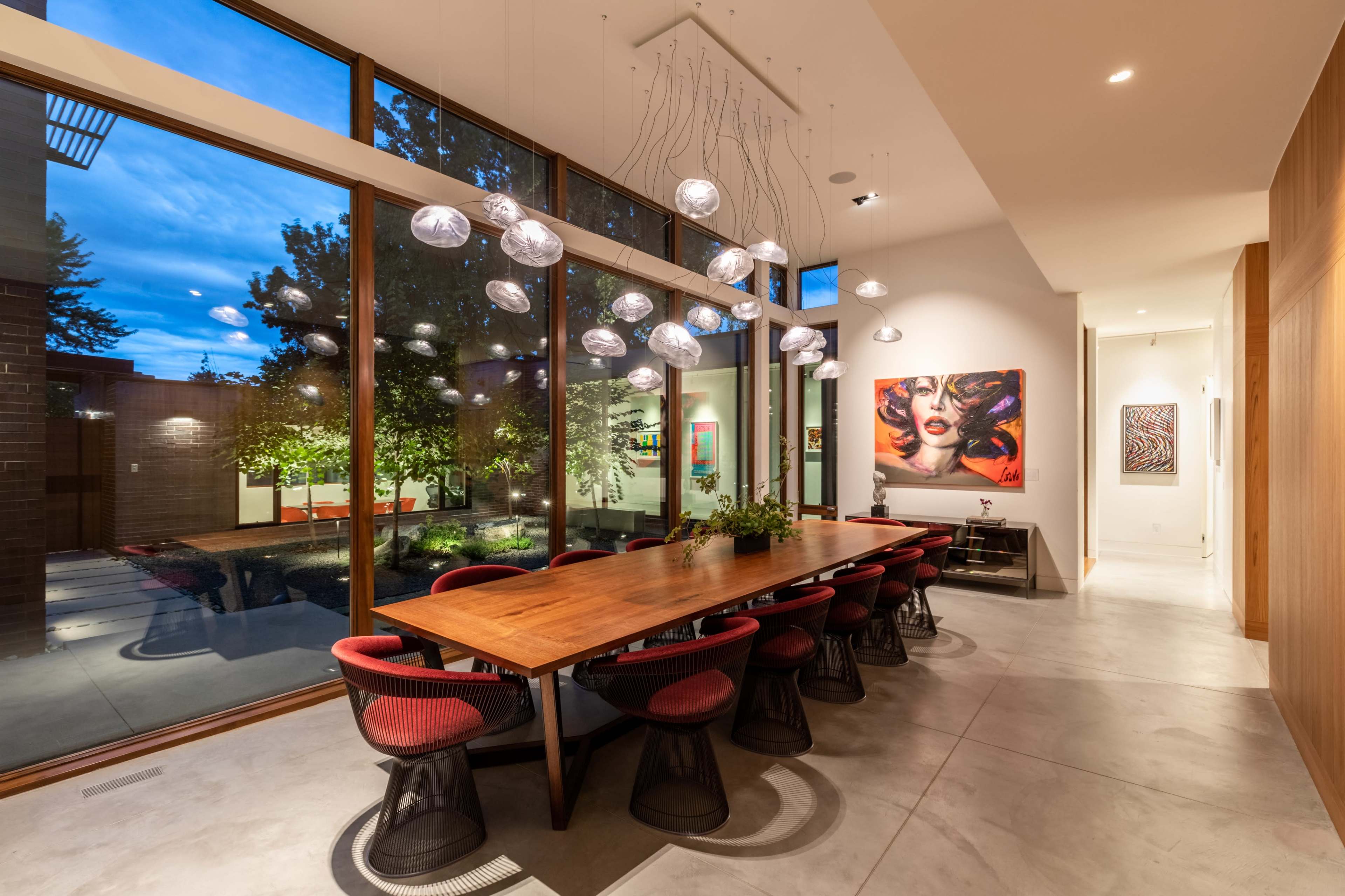 A spacious dining area features a long wooden table surrounded by modern chairs, with large windows providing views of a courtyard and artistic lighting overhead.