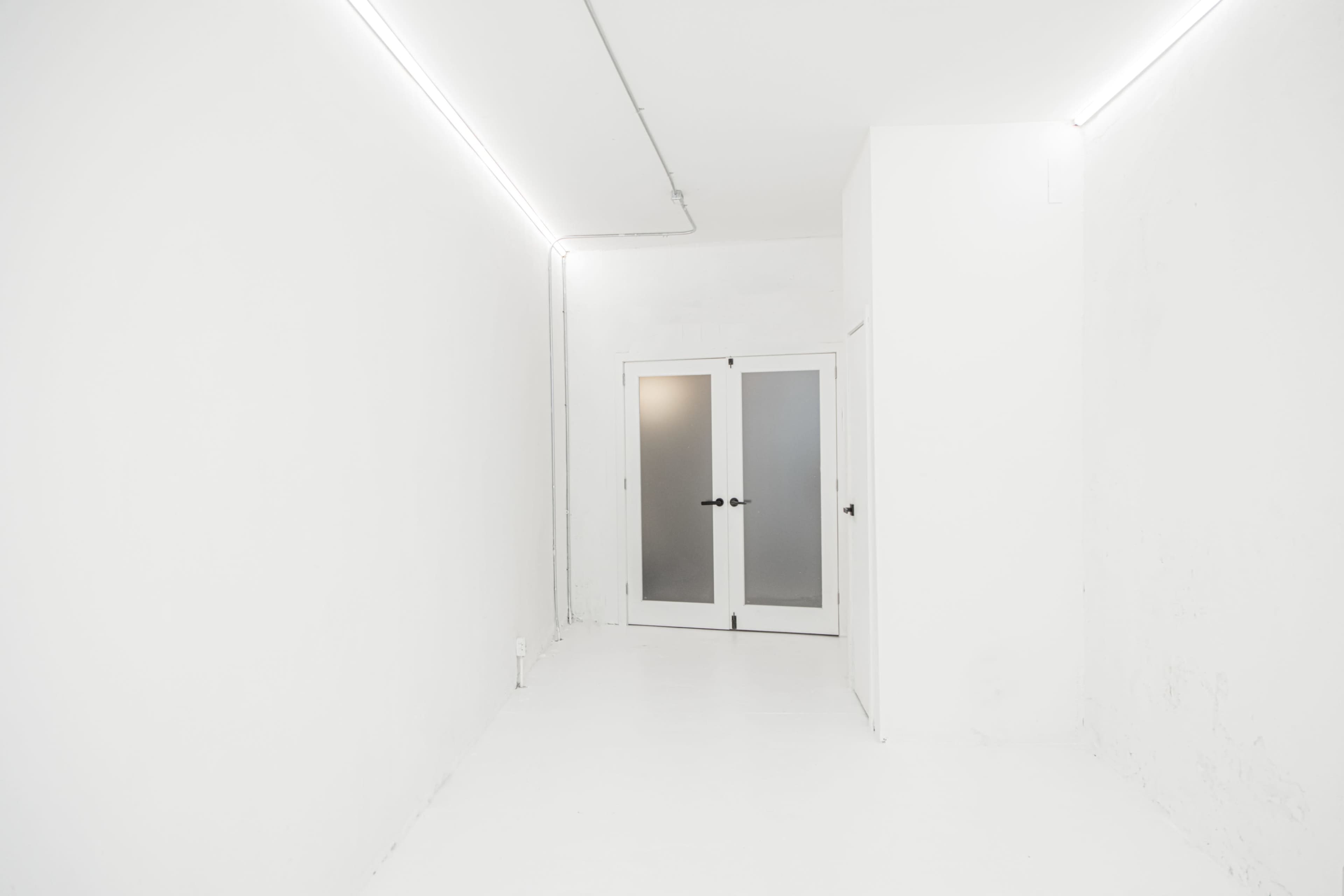 The image shows a minimalist, white hallway leading to a set of double doors with frosted glass.