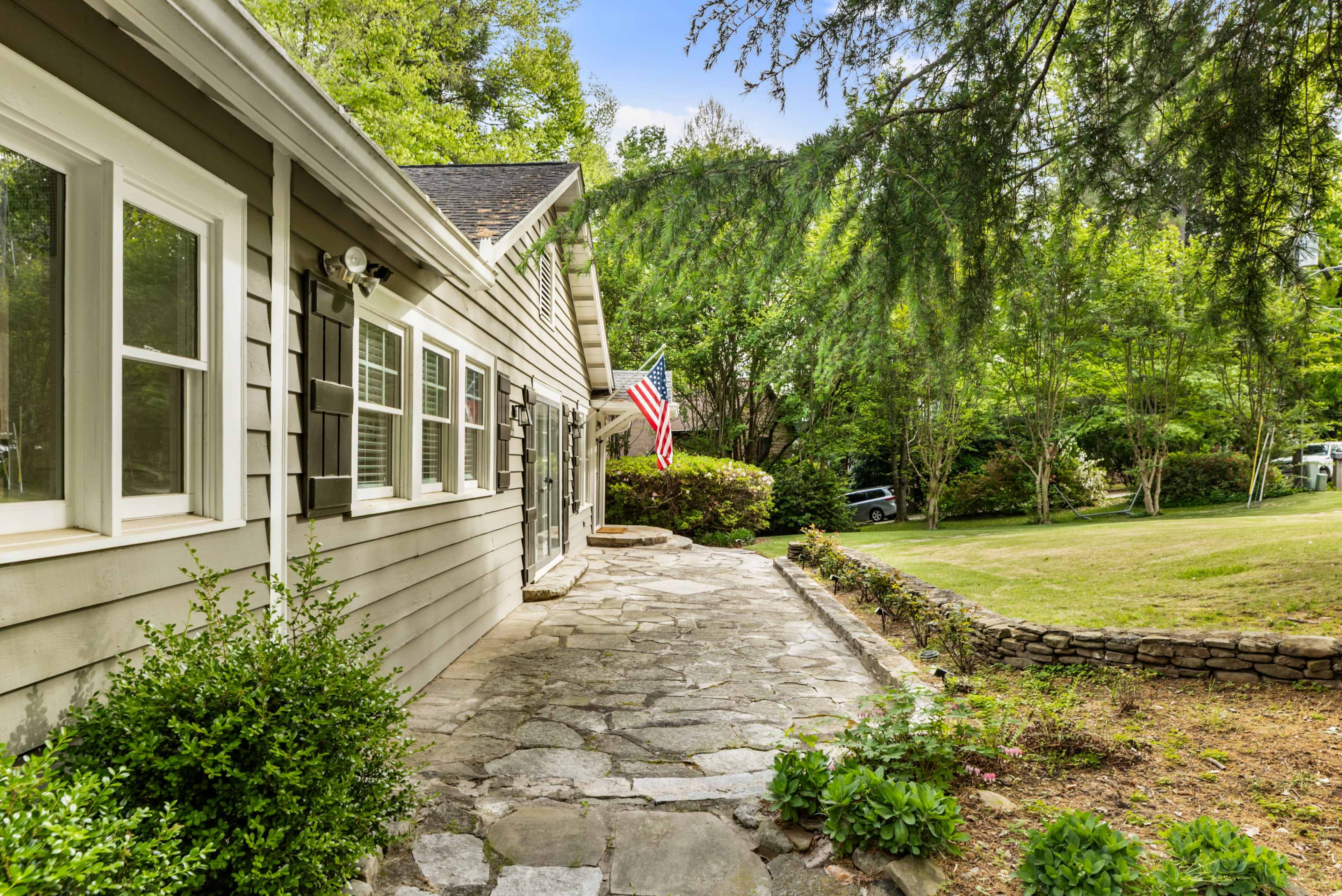 A stone path leads alongside a gray house with an American flag hanging, bordered by greenery and trees.