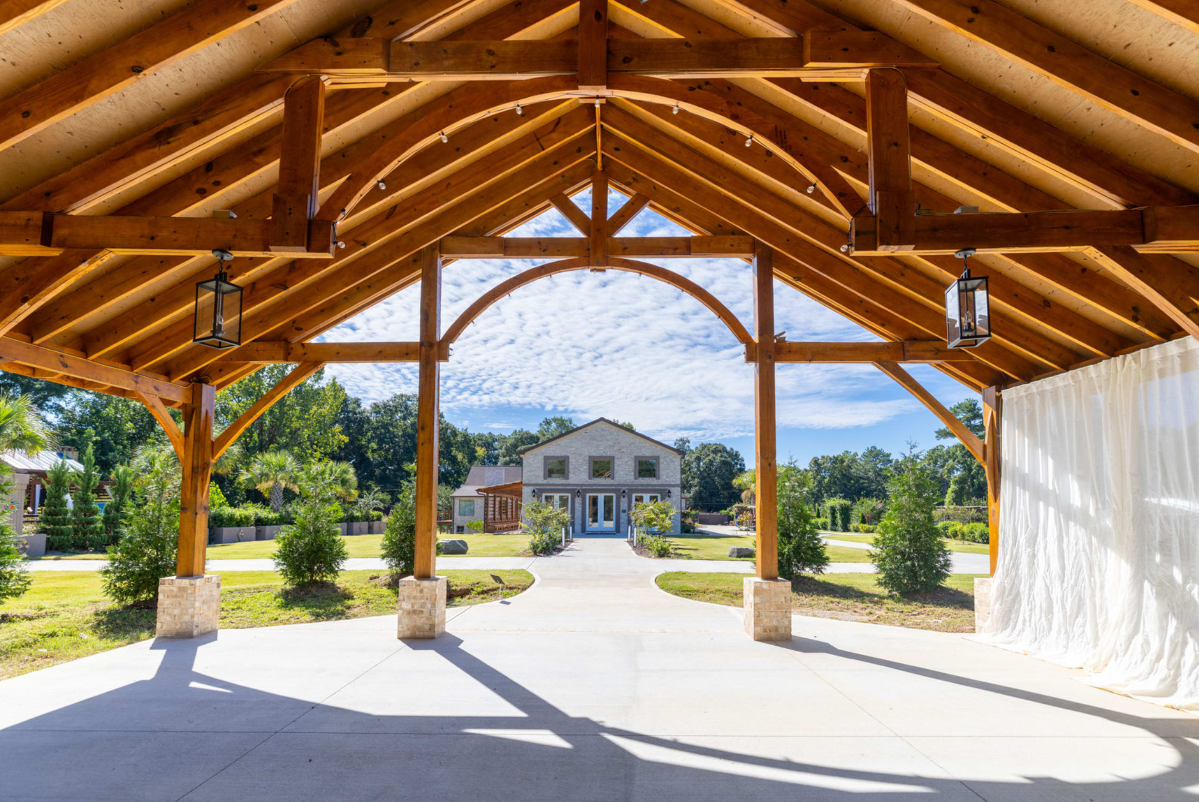 The image shows a large open structure with wooden beams, leading to a spacious lawn and a two-story building in the background.