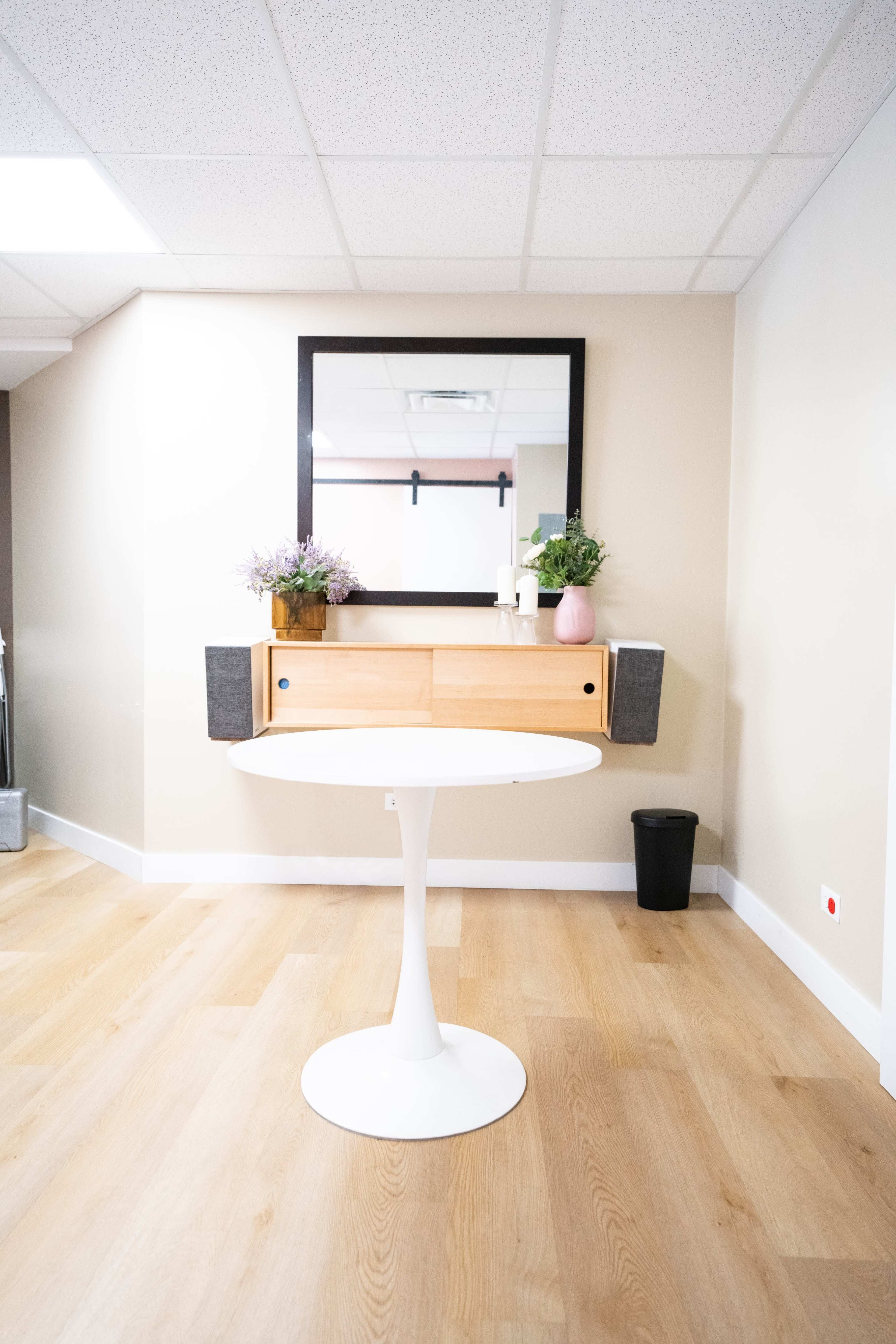 A round white dining table is centered in a room with wooden floors, featuring a wall-mounted mirror above a wooden console flanked by potted plants.