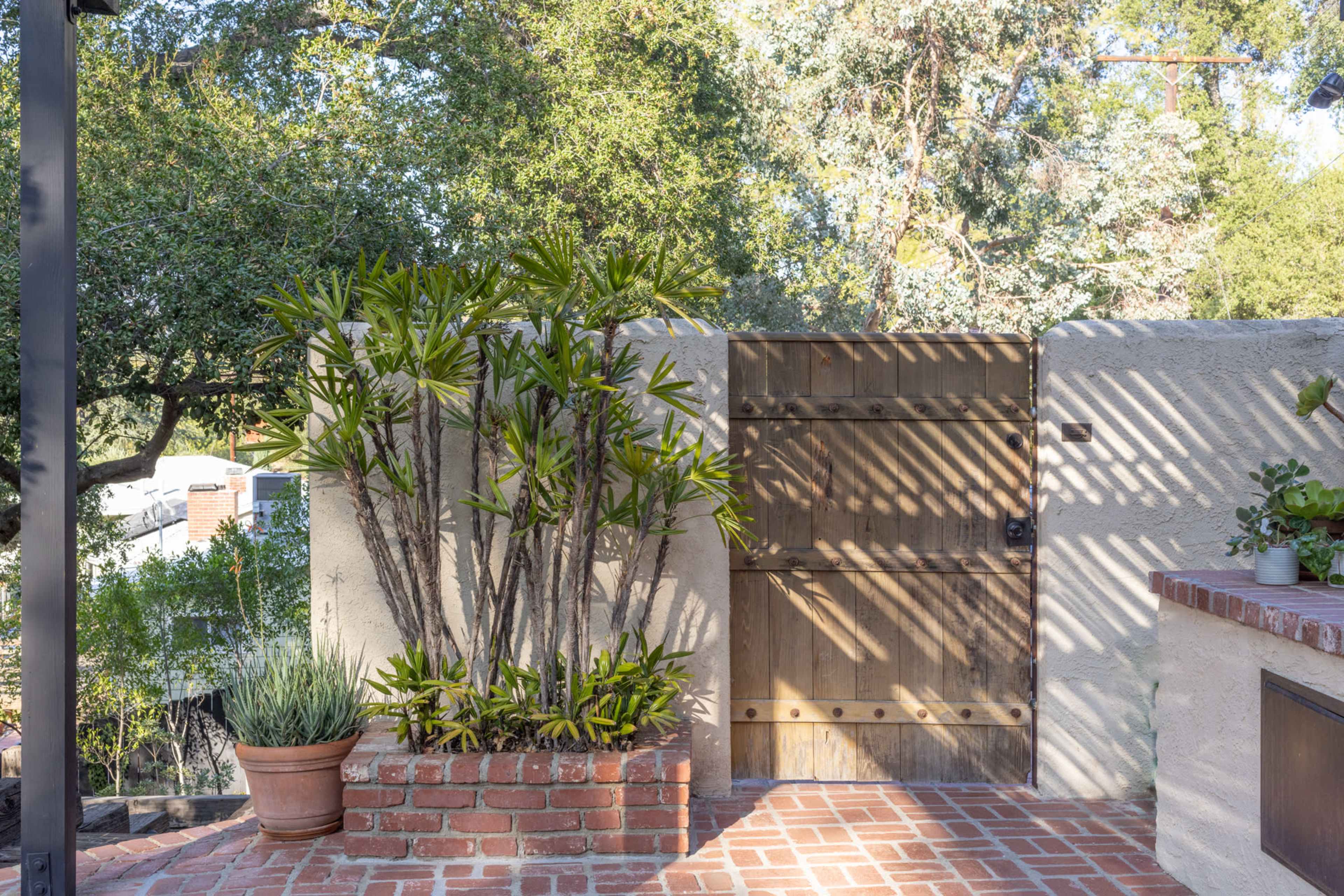 A wooden gate stands against a wall adorned with potted plants and surrounded by trees, with patterned shadows cast on the brick pathway.