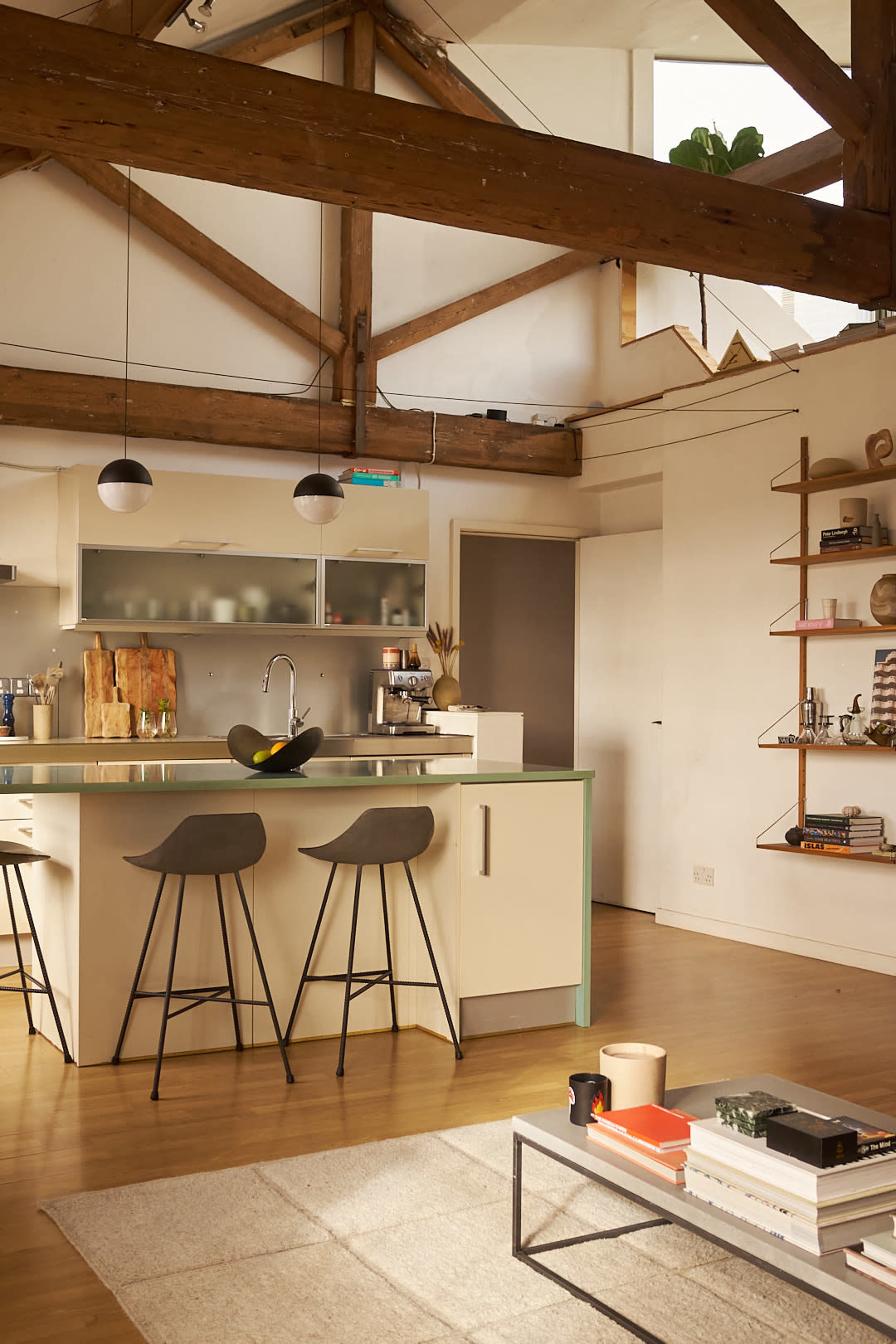 The image shows a modern kitchen with an island, two bar stools, and open shelving, all set in a spacious room featuring wooden beams.