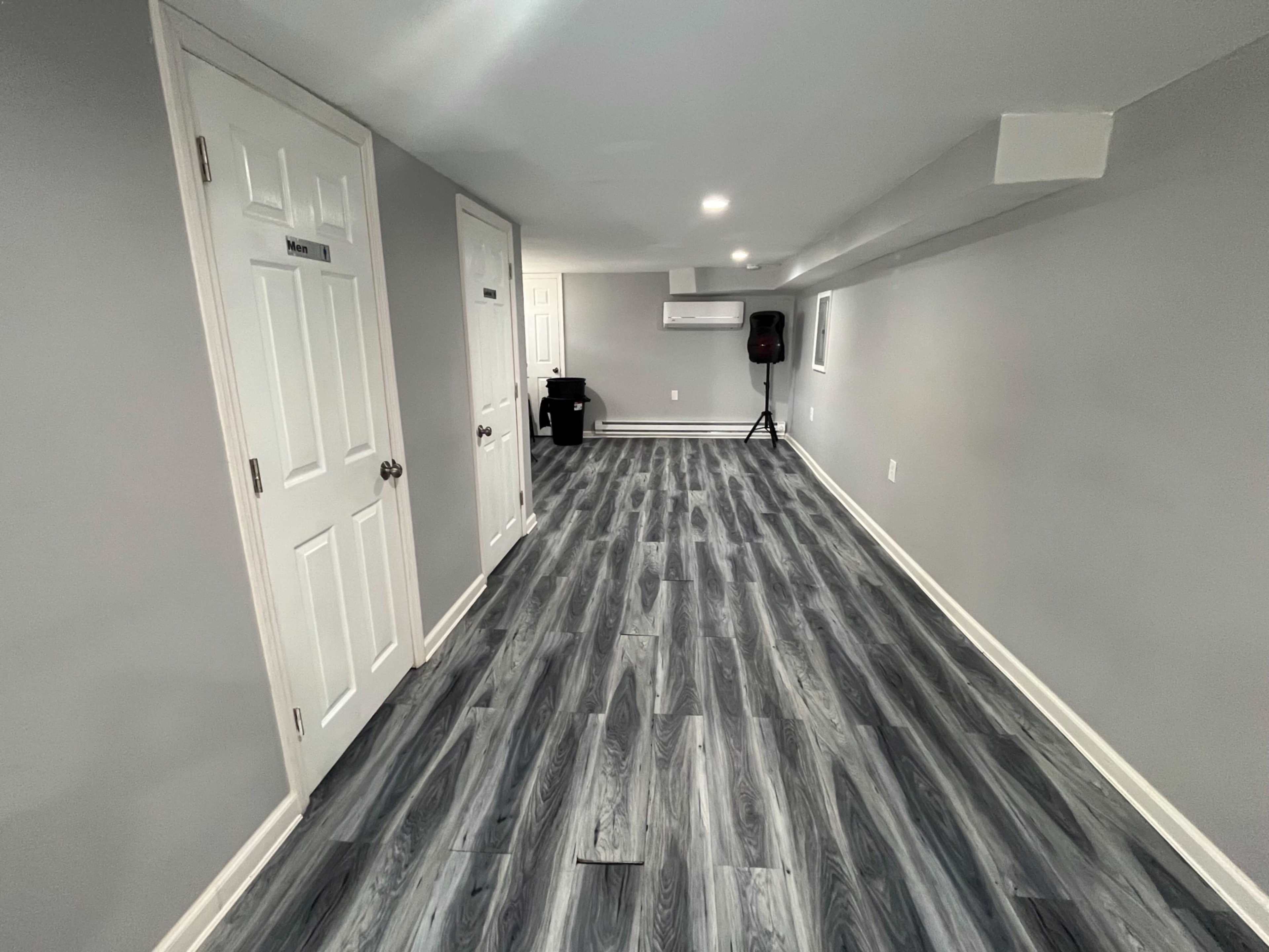 A narrow, well-lit room with gray walls and a wooden floor, featuring two closed doors on one side.
