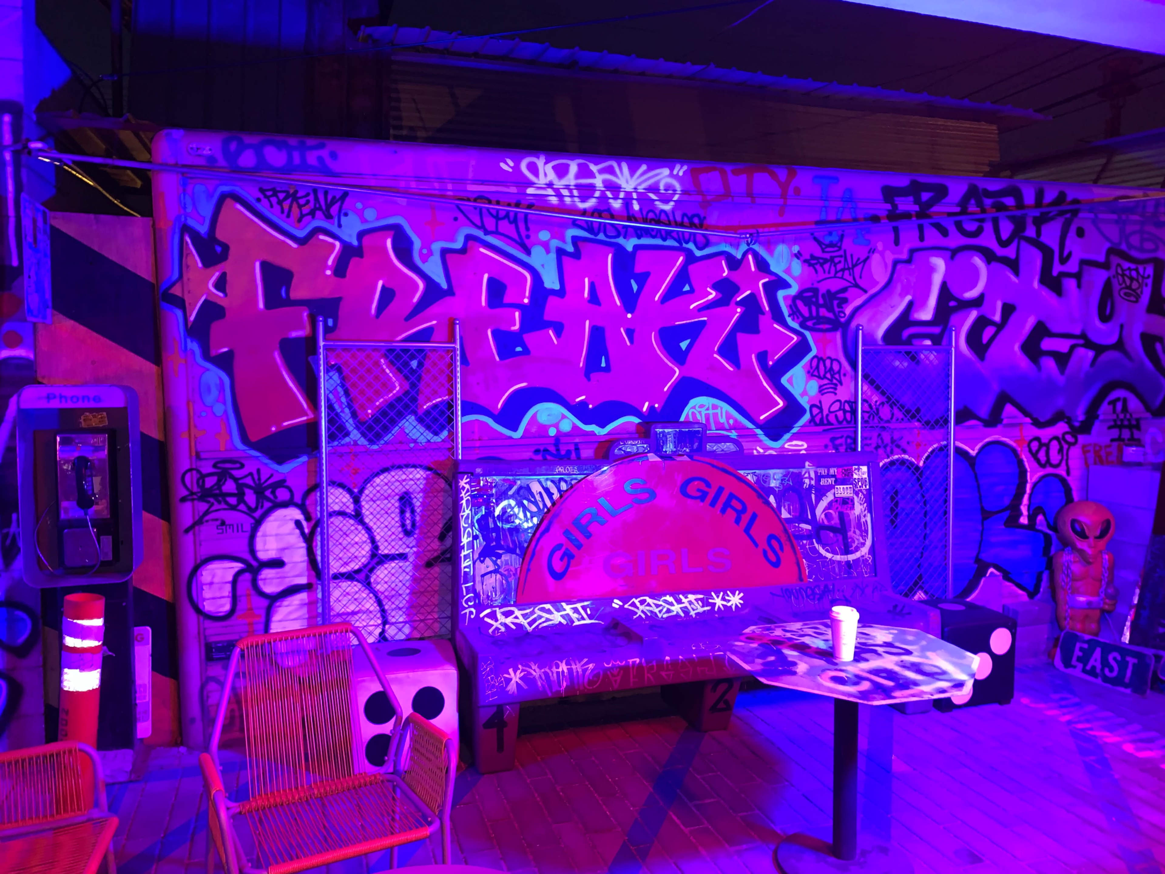 A bench labeled "GIRLS GIRLS GIRLS" is positioned in front of a colorful graffiti wall featuring various tags and designs, under purple lighting.