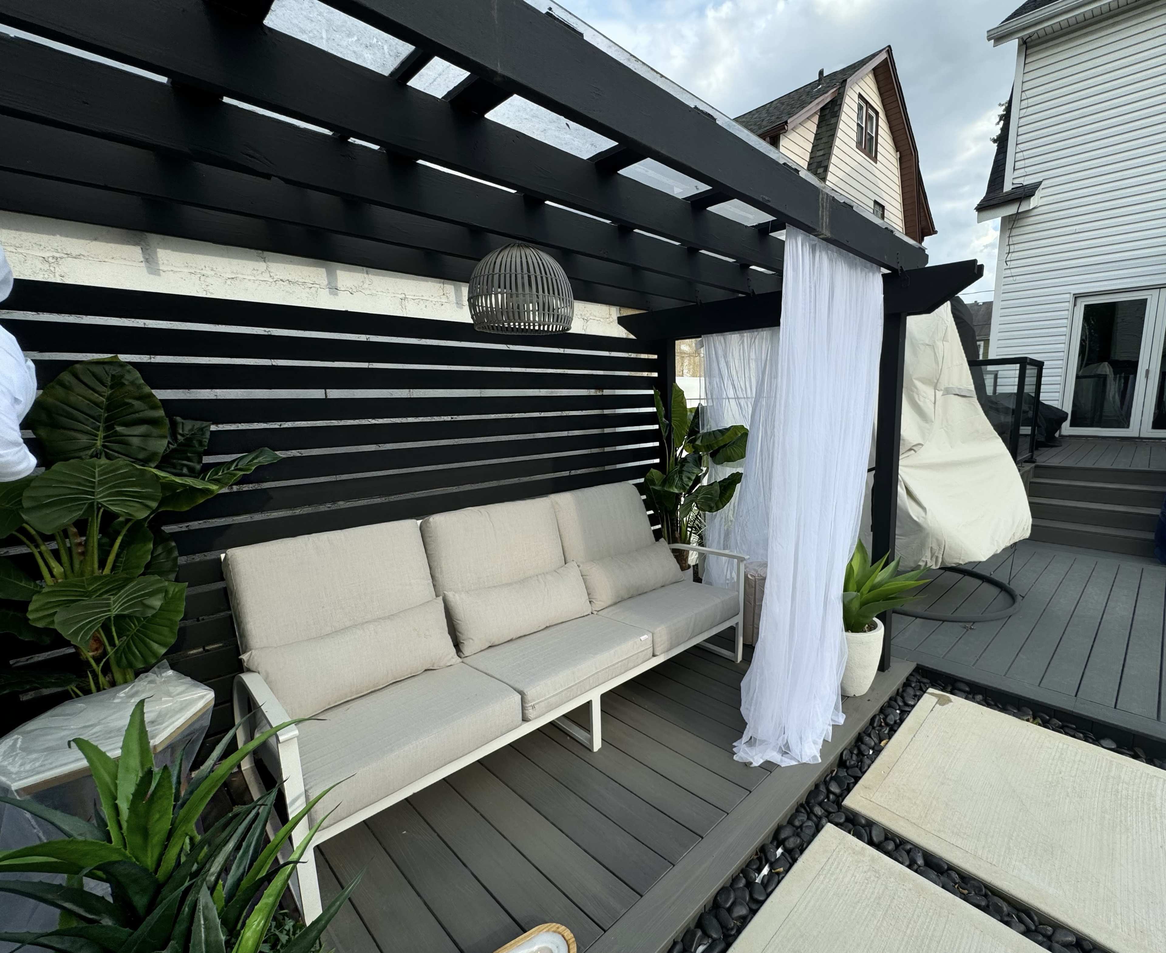 The image shows a modern outdoor seating area with a white sofa under a black pergola, complemented by potted plants and a sheer curtain.