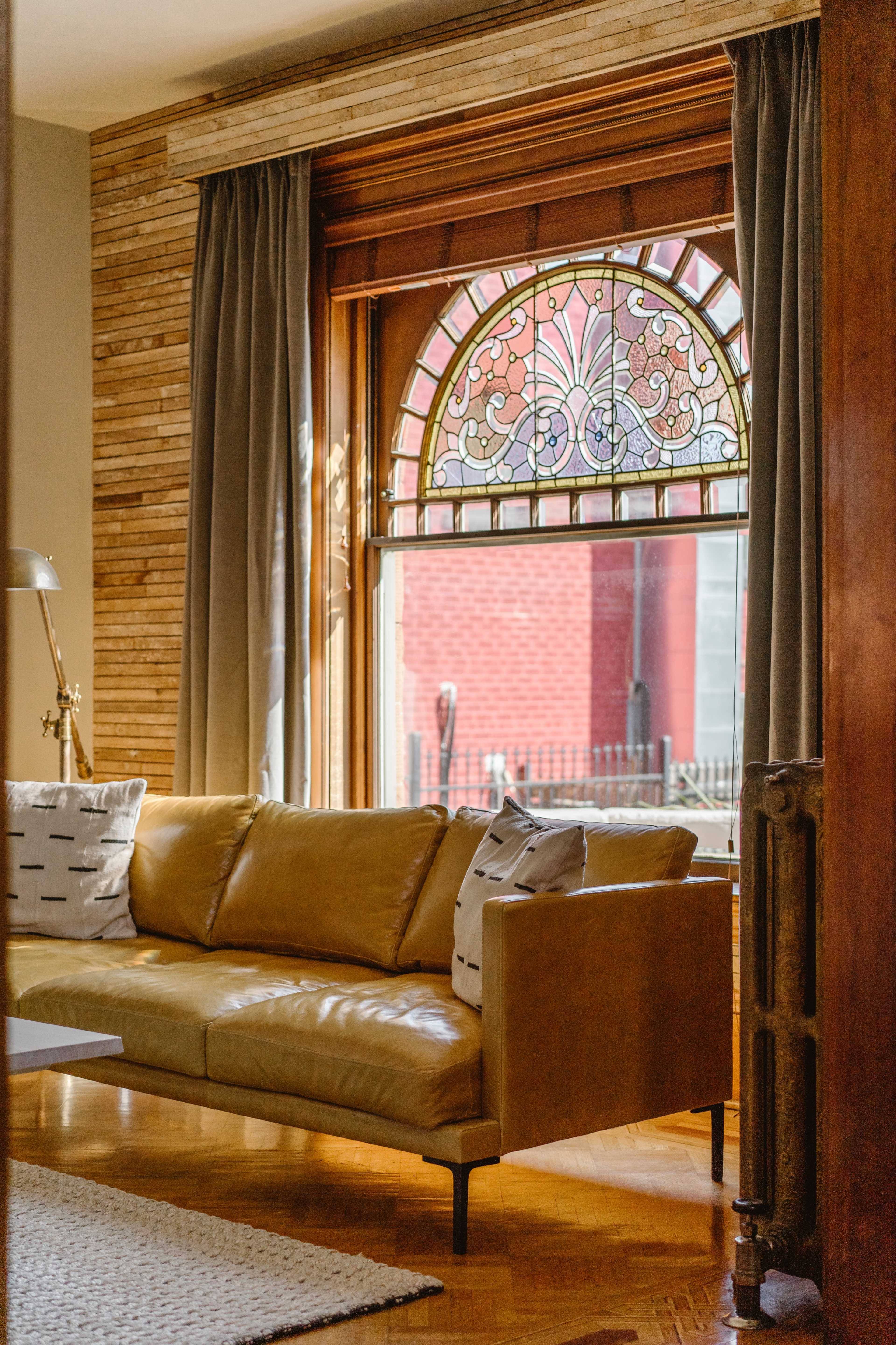 A leather sofa sits in front of a large stained glass window, framed by curtains, in a cozy living room.
