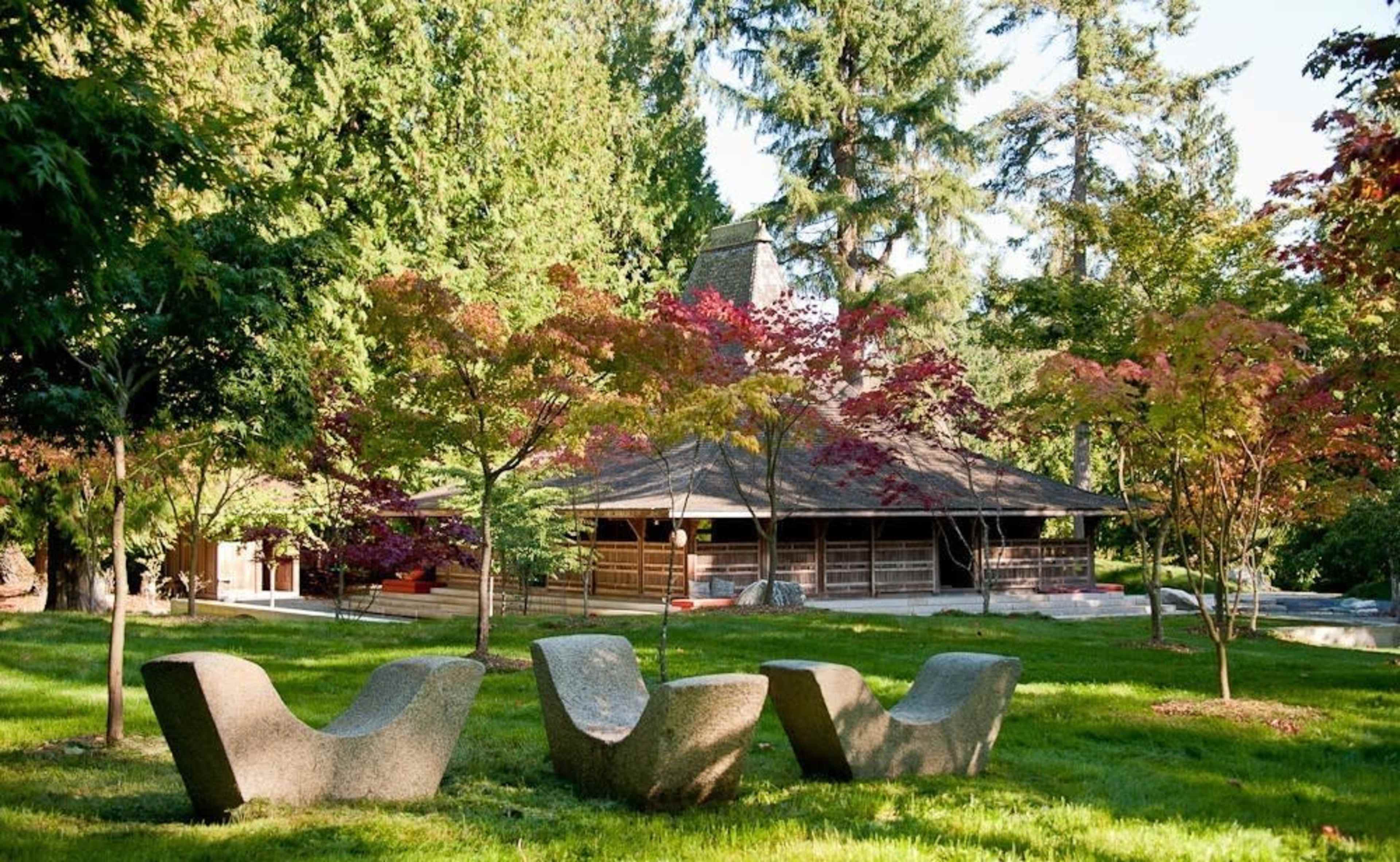The image shows a landscaped garden with vibrant trees and unique stone seating in the foreground, alongside a wooden structure in the background.