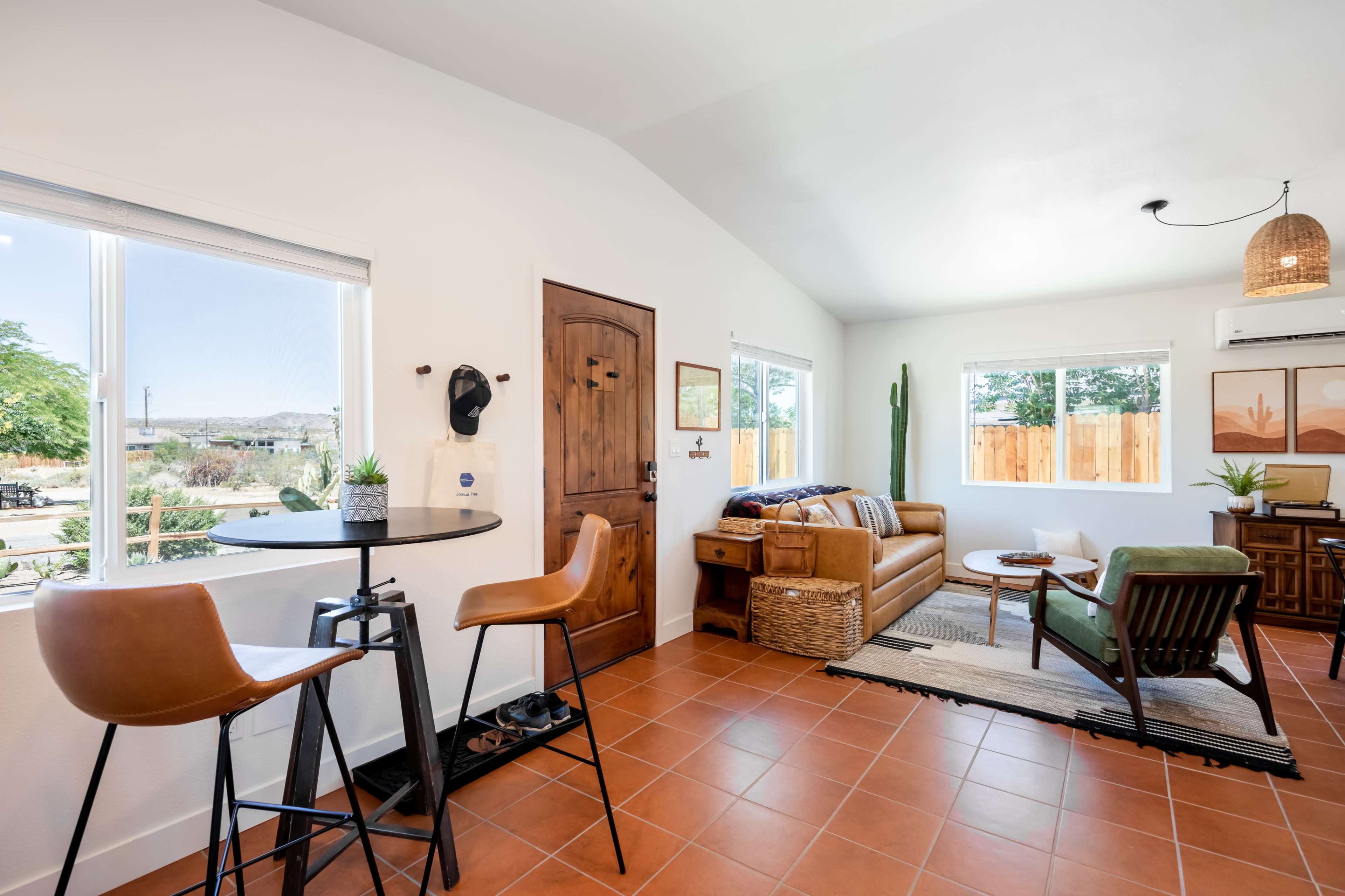 A bright, contemporary living room features a tan couch, a wooden door, and a small dining area with bar stools near a window showcasing a scenic outdoor view.