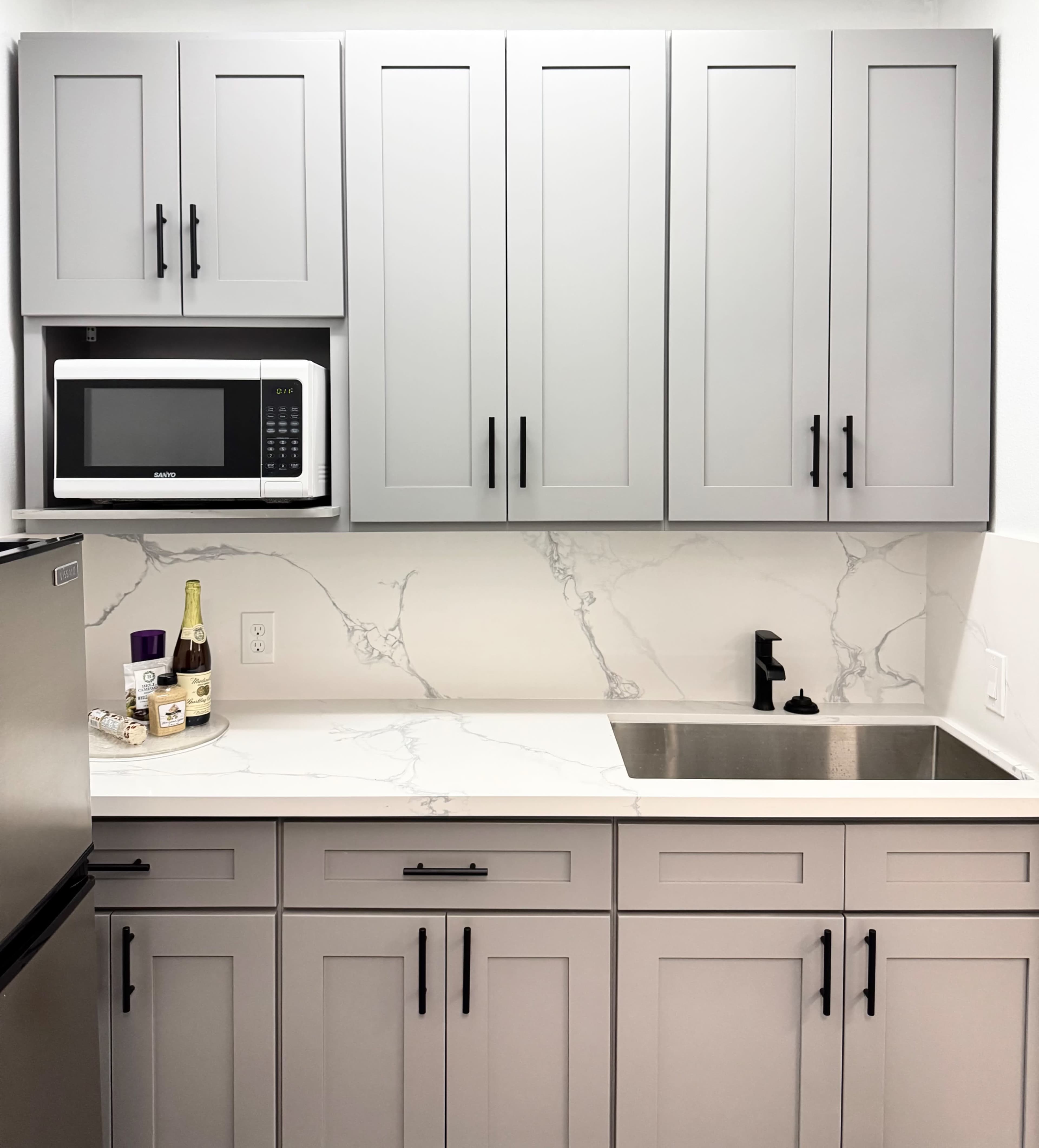 The image shows a modern kitchen with gray cabinets, a microwave above the sink, and a marble countertop featuring a sink and a bottle of wine.