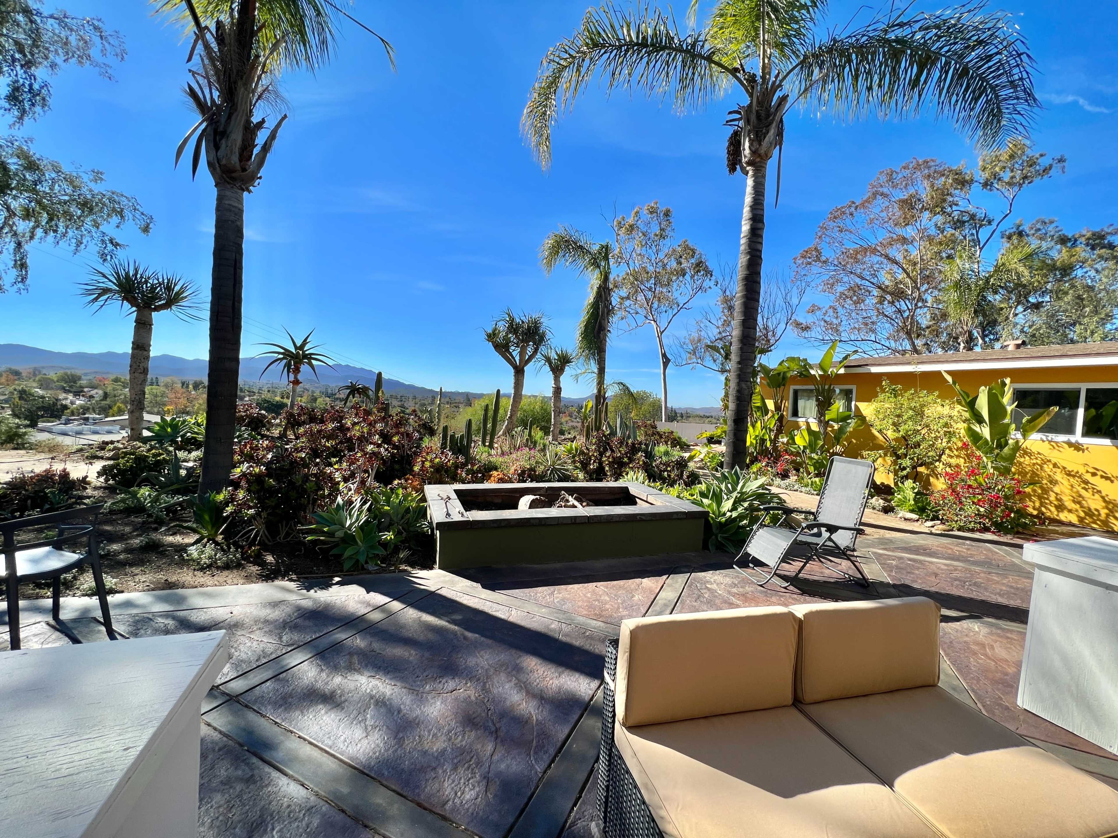 A landscaped outdoor area featuring palm trees, colorful plants, and a fire pit, with a view of distant mountains under a clear blue sky.