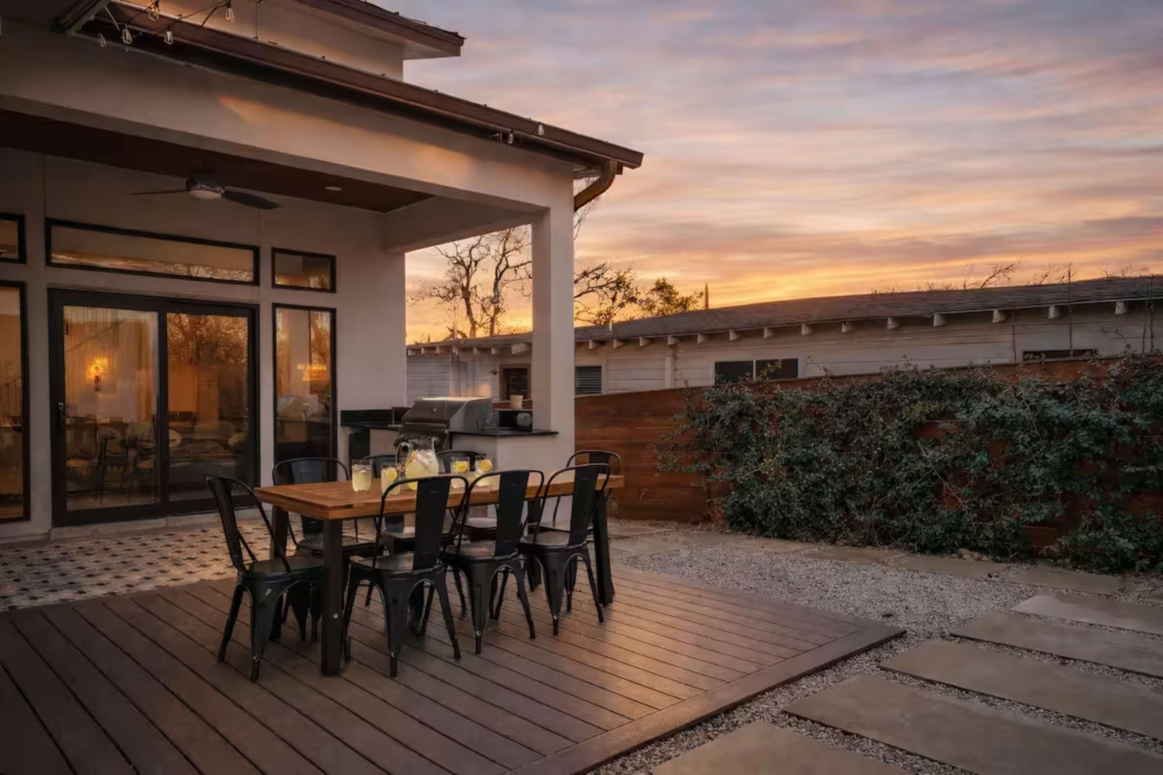 A patio area with a wooden dining table and black chairs is set against a backdrop of a sunset sky, adjacent to a house with a grill and surrounding greenery.