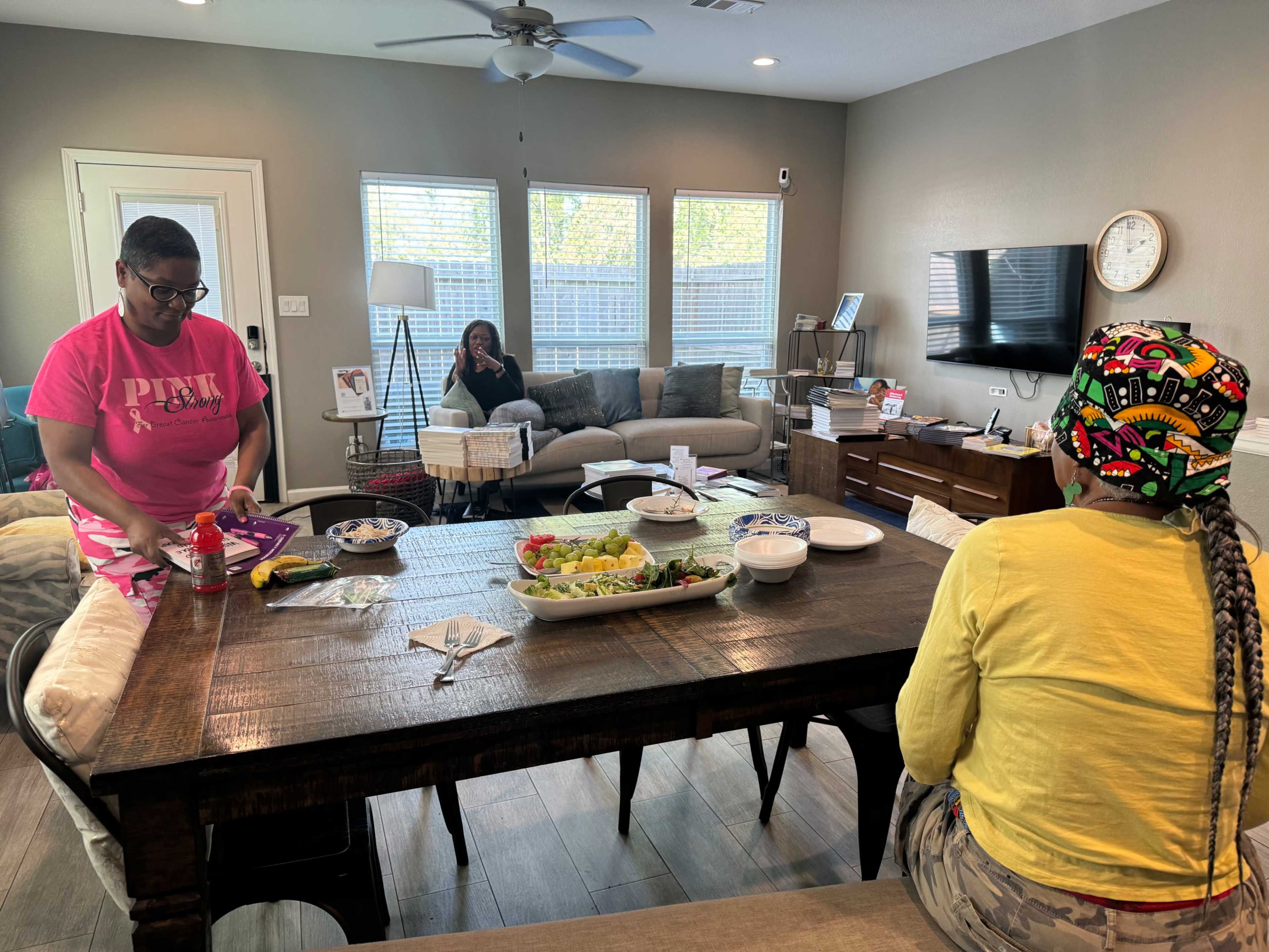 Two women are seated at a dining table, one preparing food while the other observes, with a third woman in the background relaxing on a couch in a well-lit living room.