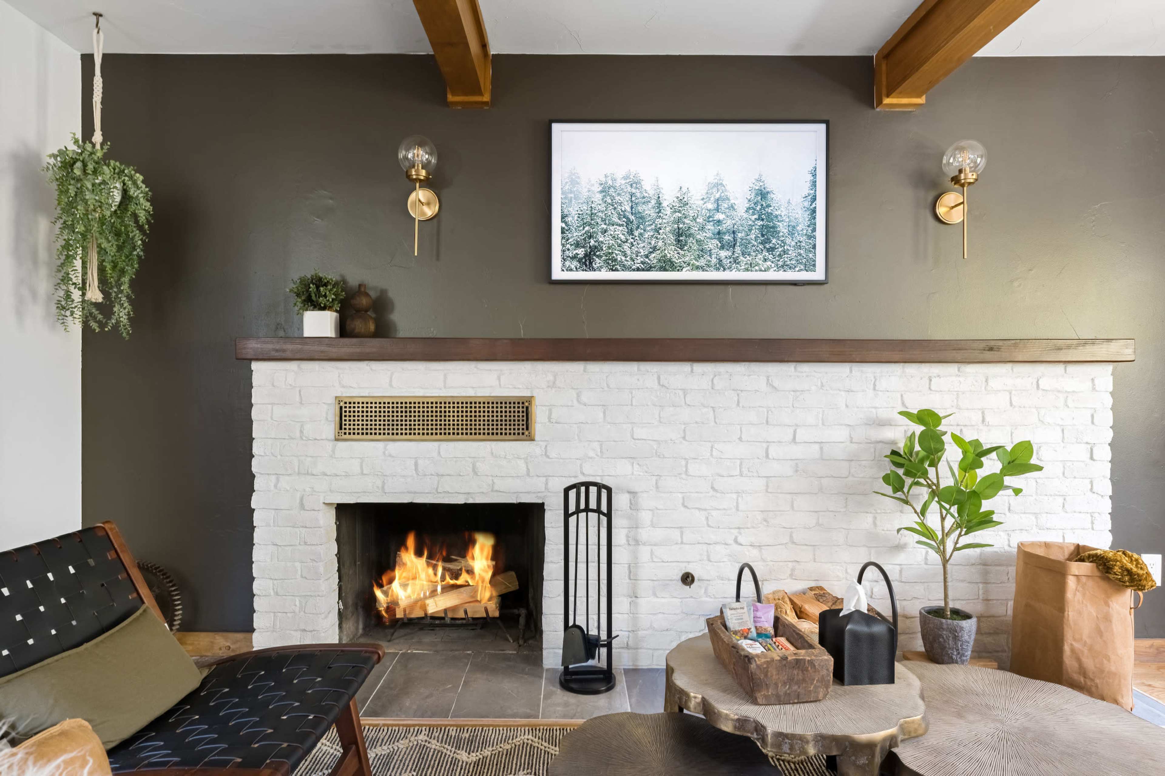 A cozy living room features a white brick fireplace with a fire burning, surrounded by modern decor elements including a potted plant and wall-mounted light fixtures.