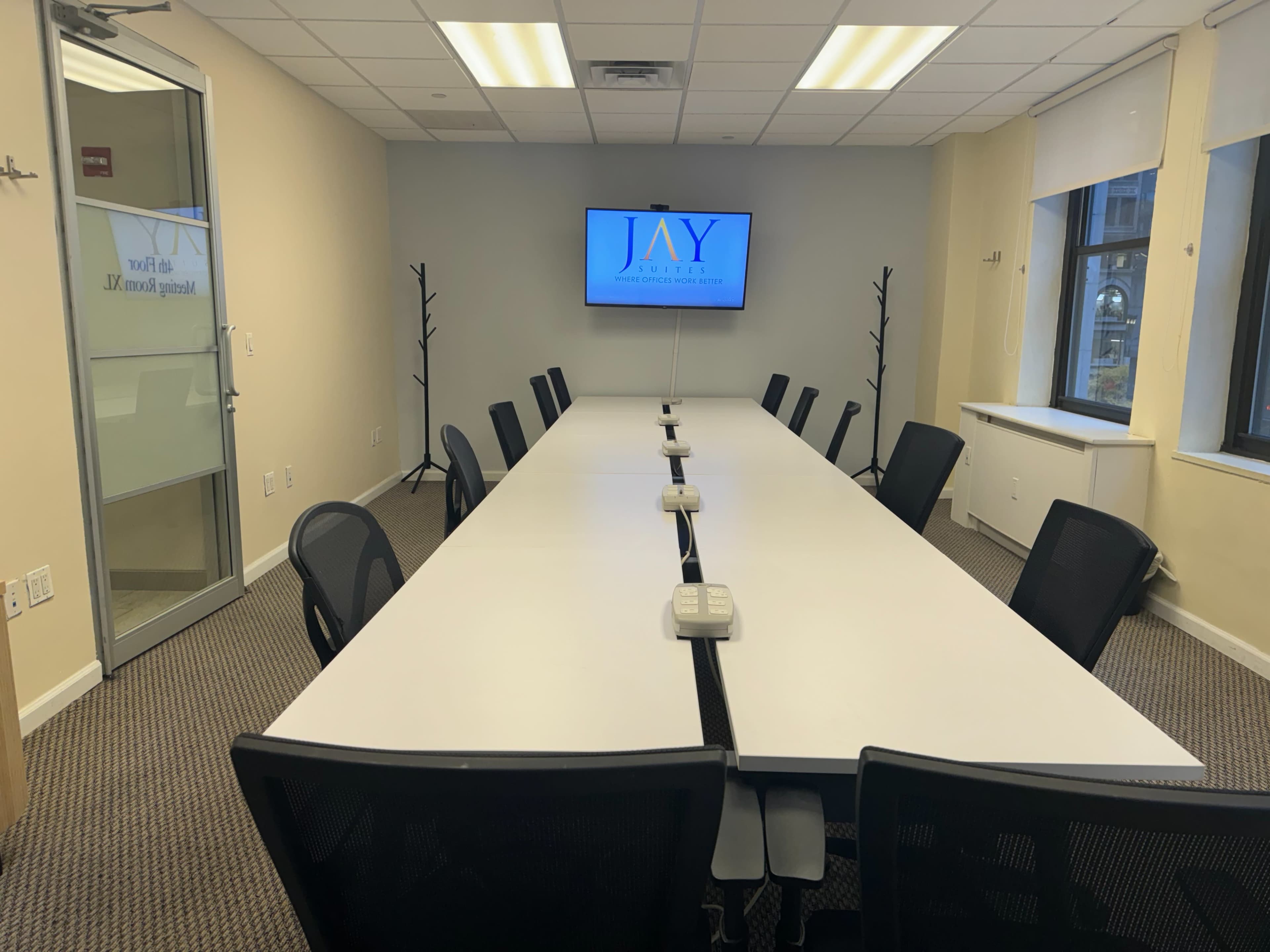 A conference room features a long white table surrounded by black chairs, with a large monitor displaying the name "JAY" on the wall.