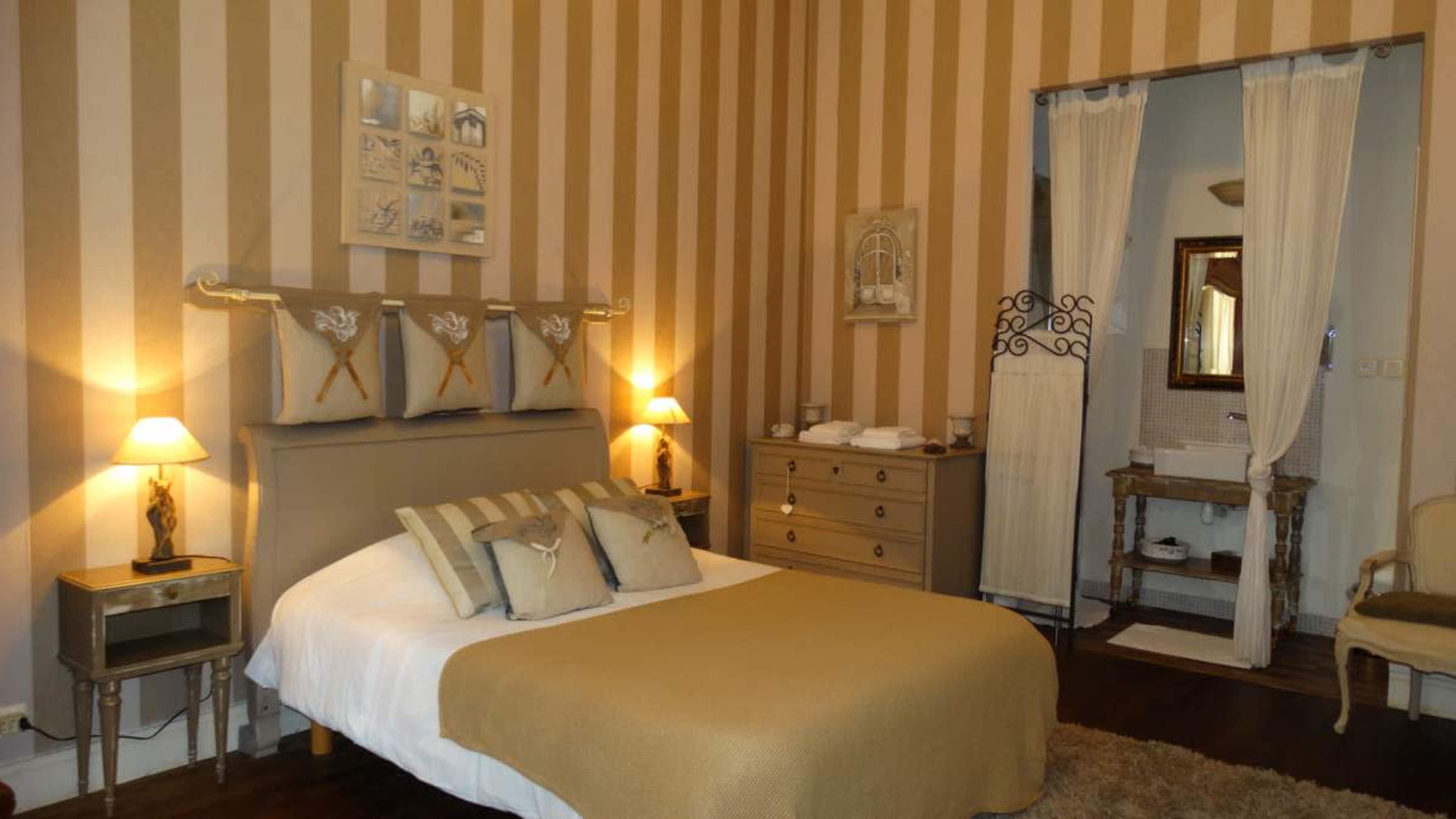 The image shows a cozy bedroom with striped walls, a bed covered with a tan blanket, and a small en-suite bathroom visible through an open door.