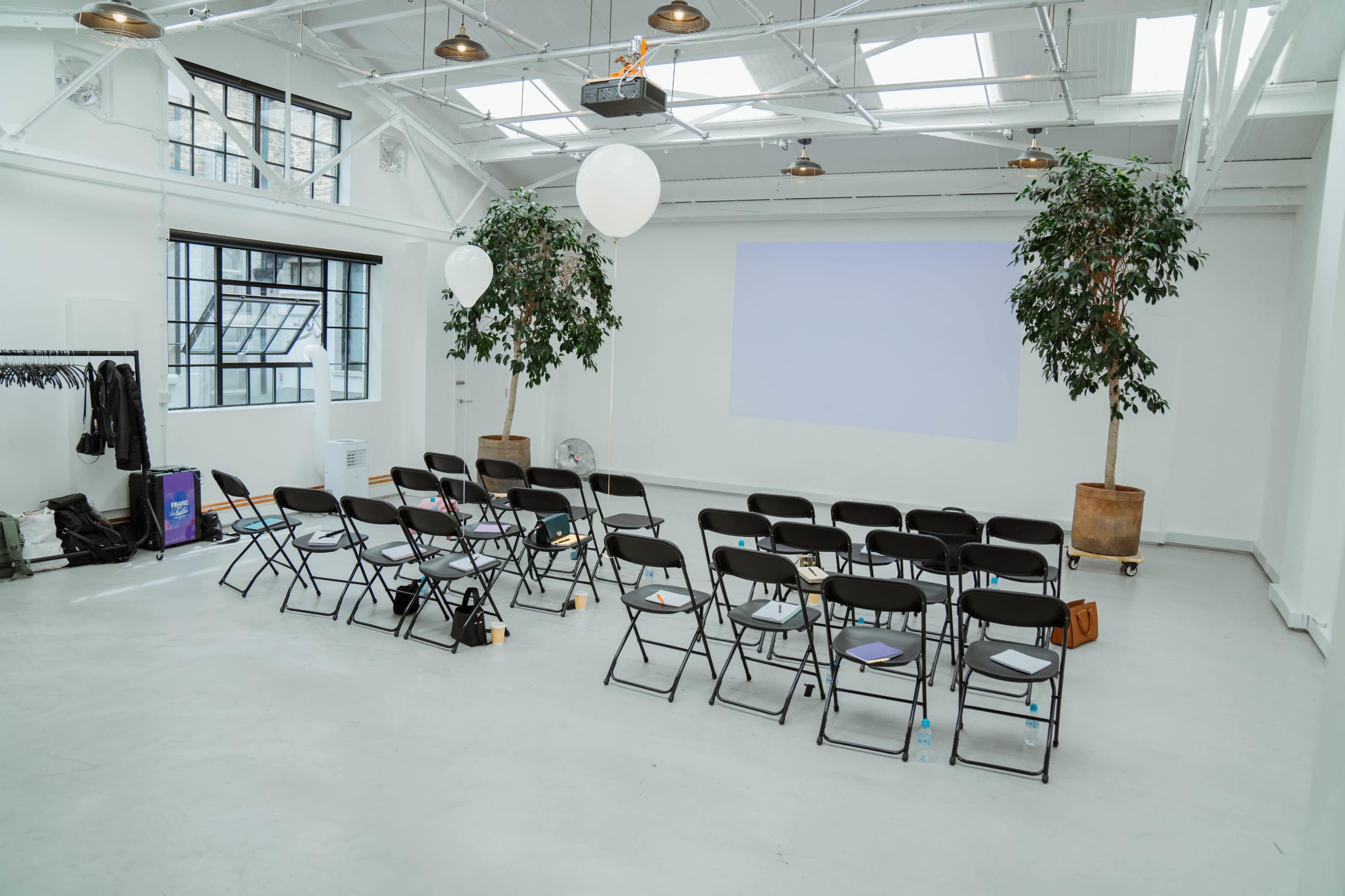A spacious, minimalist room features rows of black folding chairs facing a large blank screen, flanked by two potted trees and decorated with white balloons.