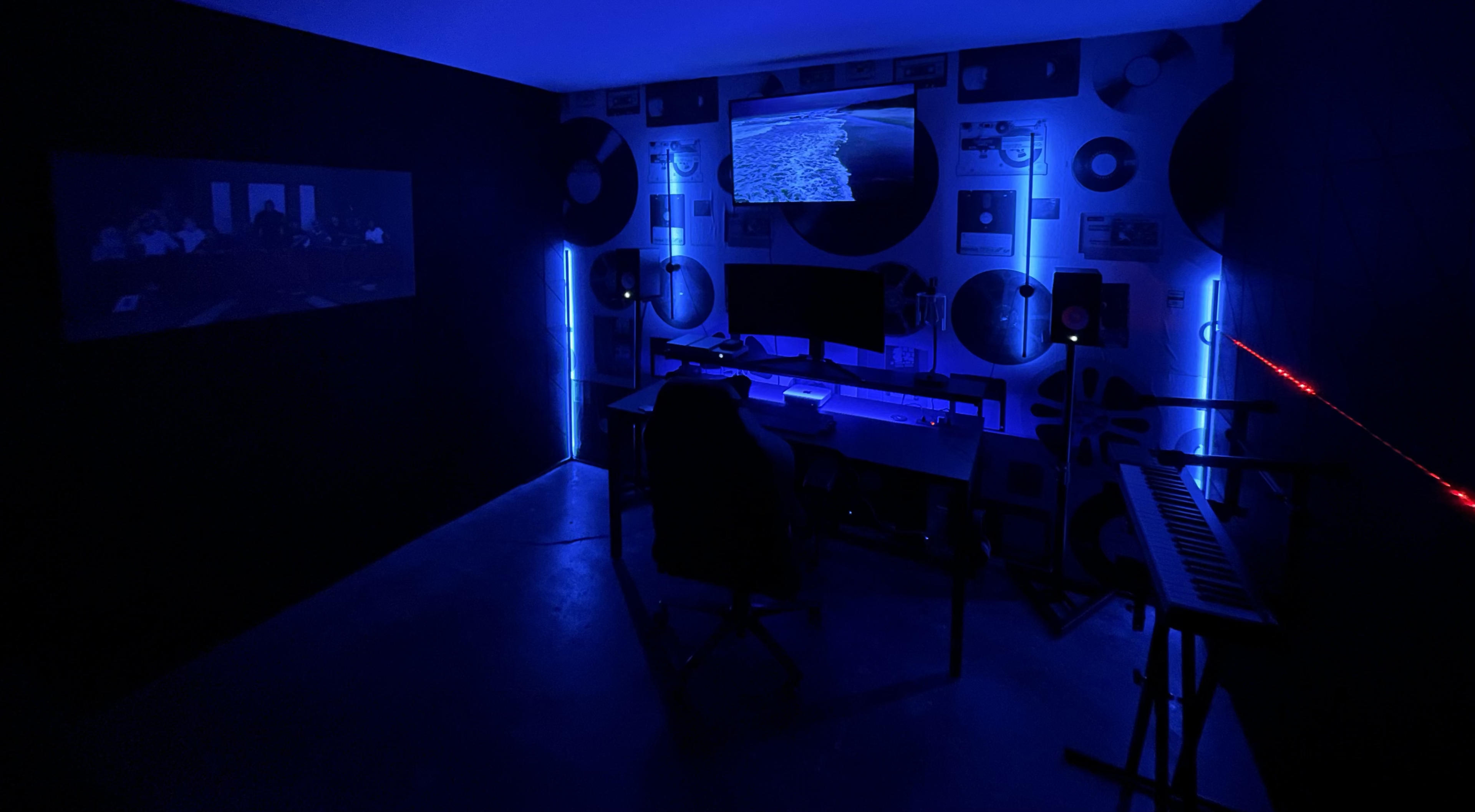 A dimly lit room features a desk with a computer monitor surrounded by blue LED lights, a keyboard, and a wall adorned with circular decor.