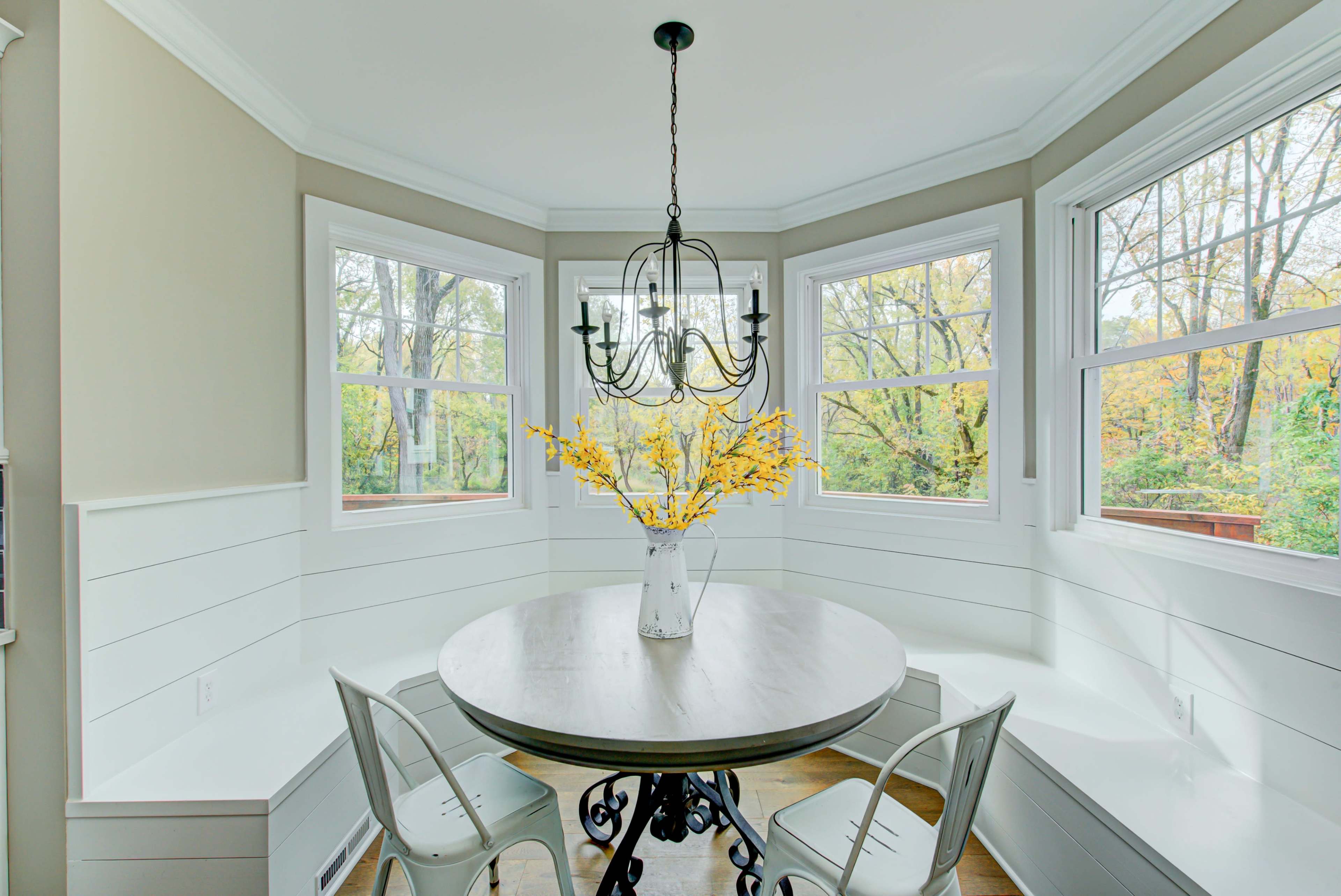 A round table with a vase of yellow flowers sits in a bright corner nook surrounded by windows.