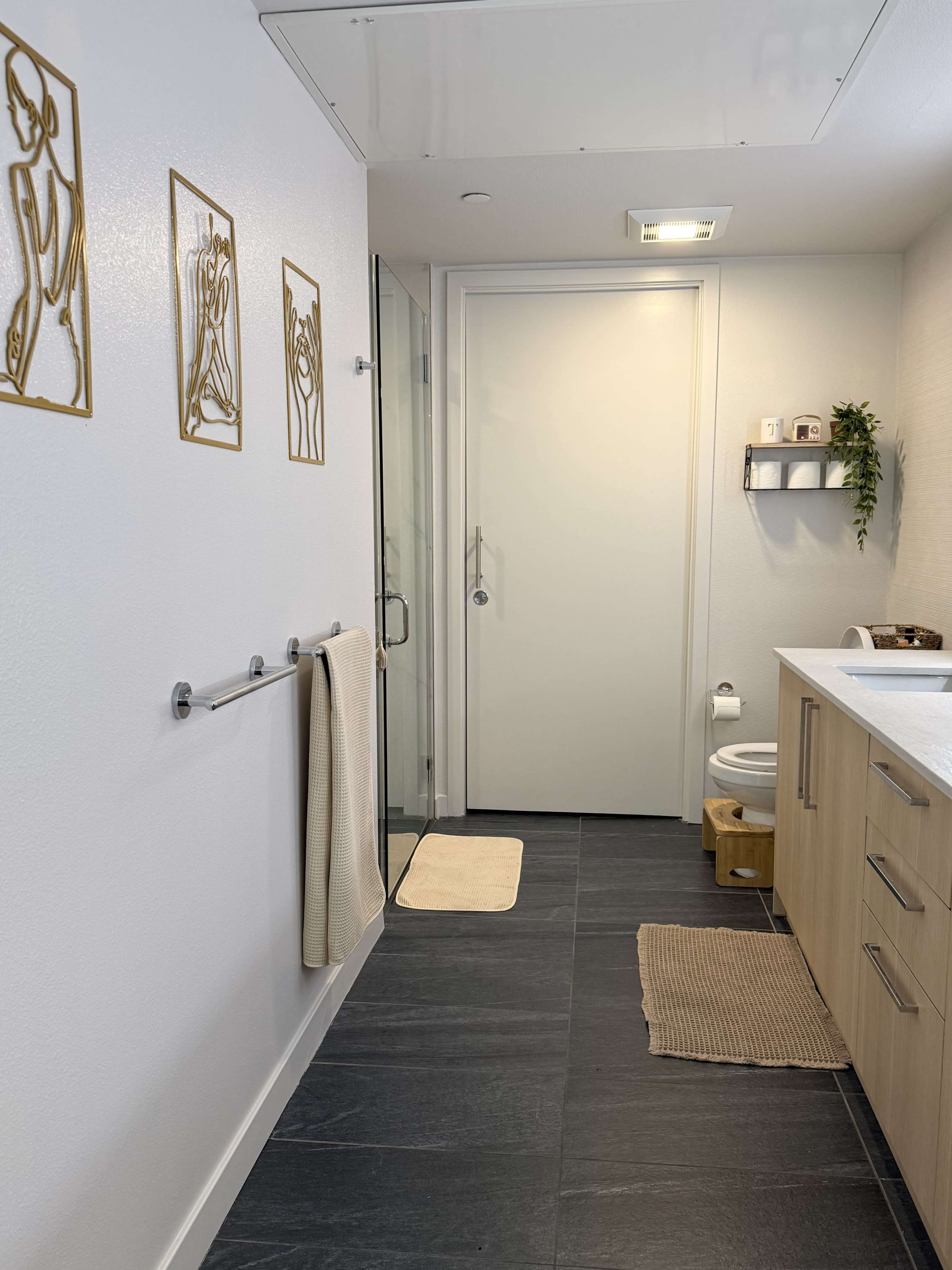 The image shows a modern bathroom with glass shower doors, a wooden vanity, a towel rack, and stylized wall art.