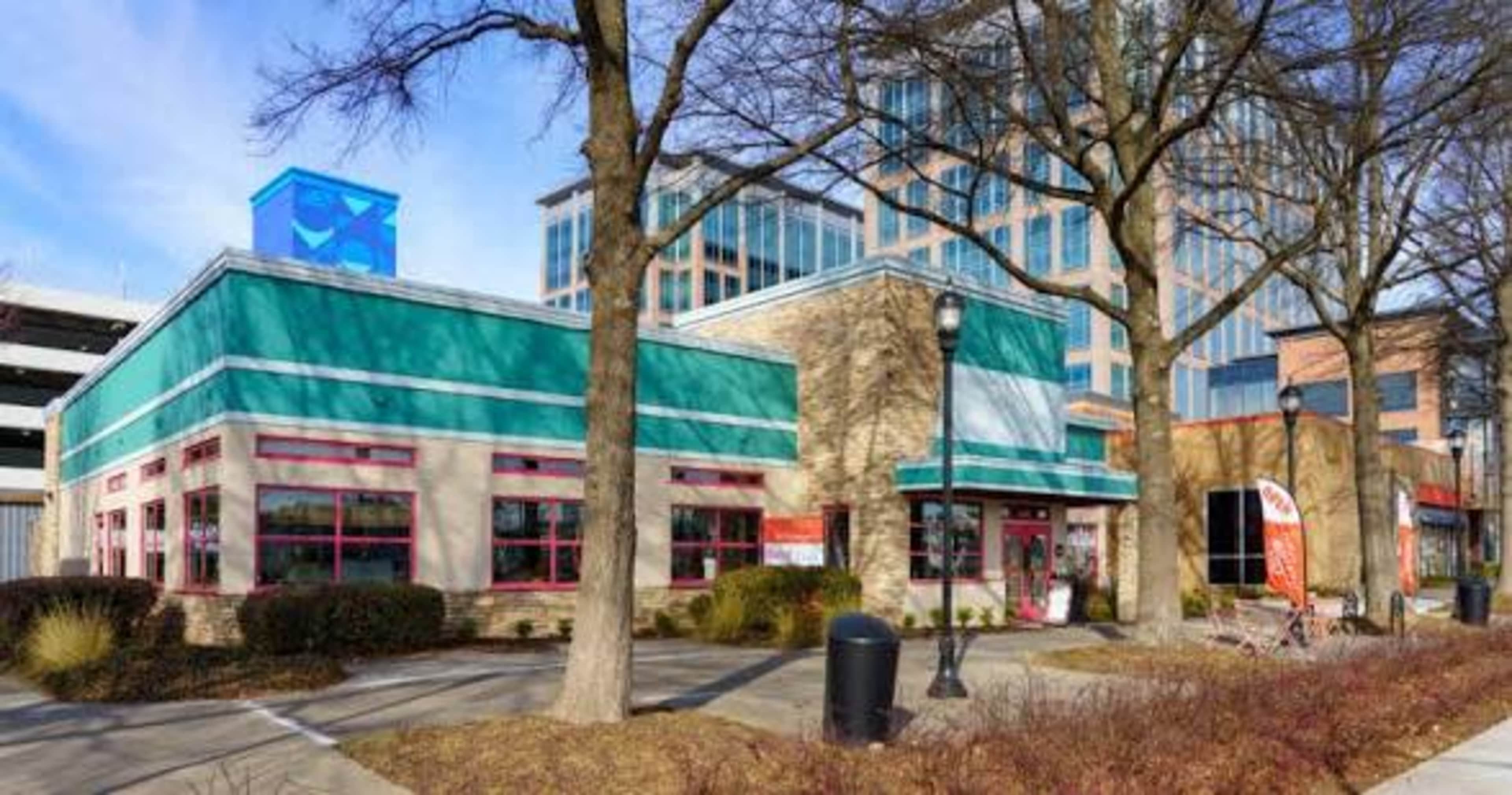 The image shows a commercial building with a vibrant turquoise and white exterior, surrounded by trees and a parking area, adjacent to a larger glass office structure.