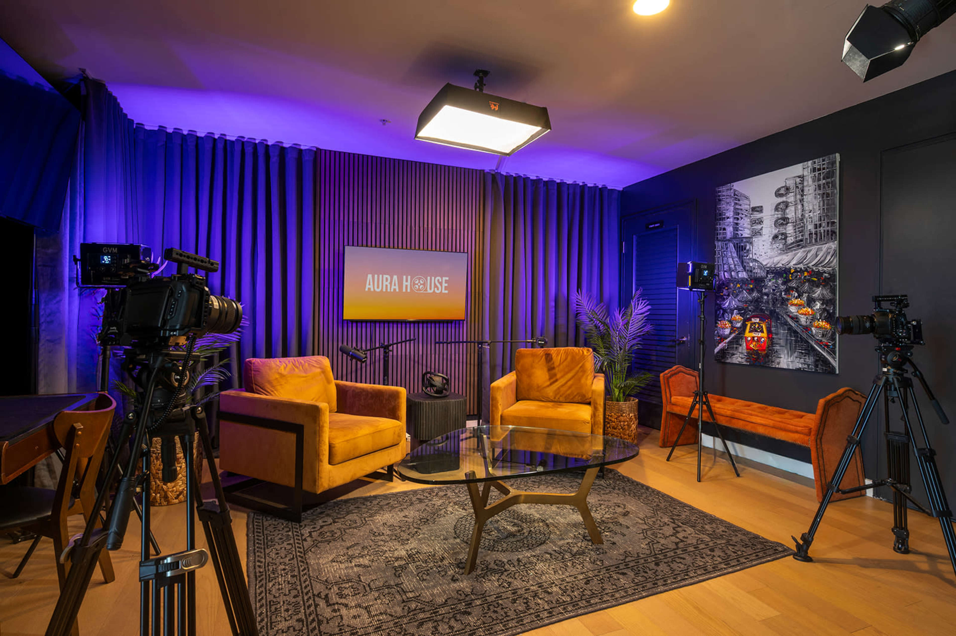 The image shows a modern studio setup with two yellow armchairs, a glass coffee table, and several cameras positioned around a backdrop featuring the words "AURA HOUSE."
