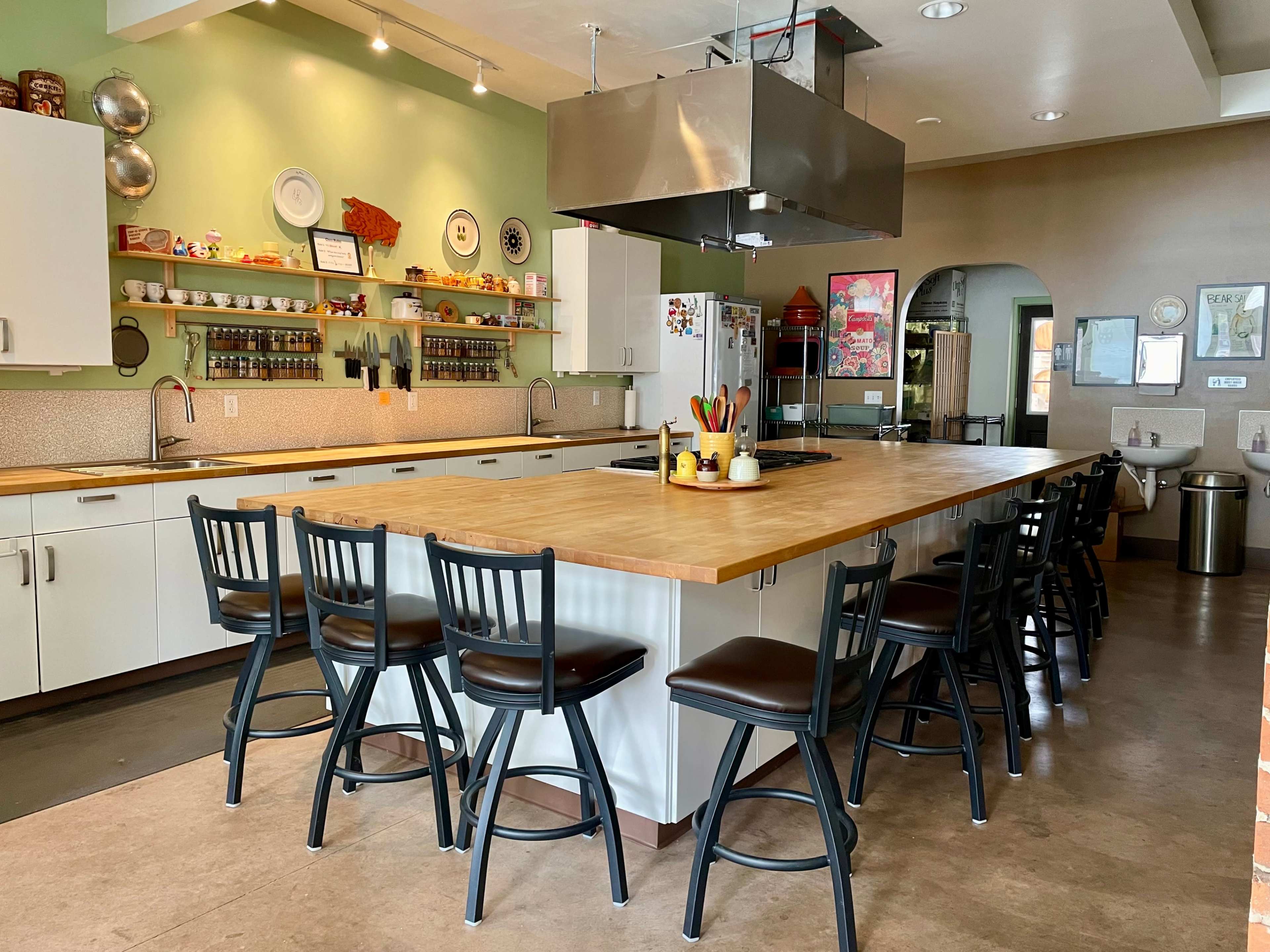 A spacious kitchen with a large wooden island surrounded by black bar stools, featuring shelves of ingredients and utensils along the walls.