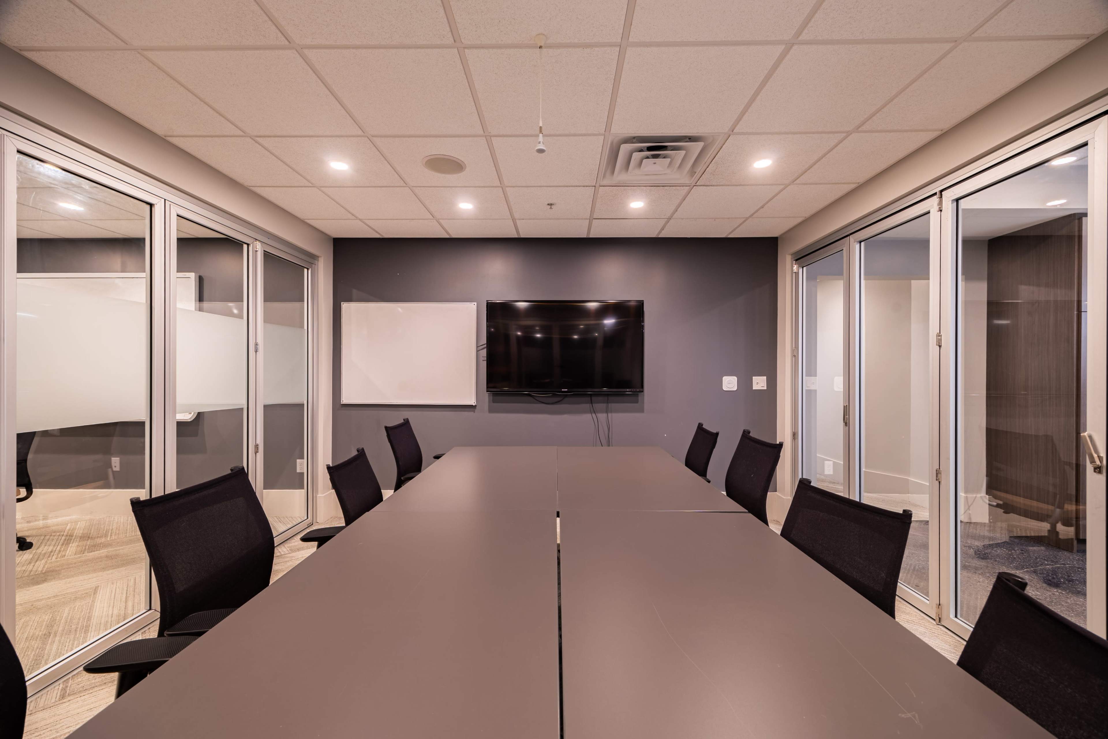 The image shows a modern conference room with a long table, black chairs, a television mounted on the wall, and glass partitions.
