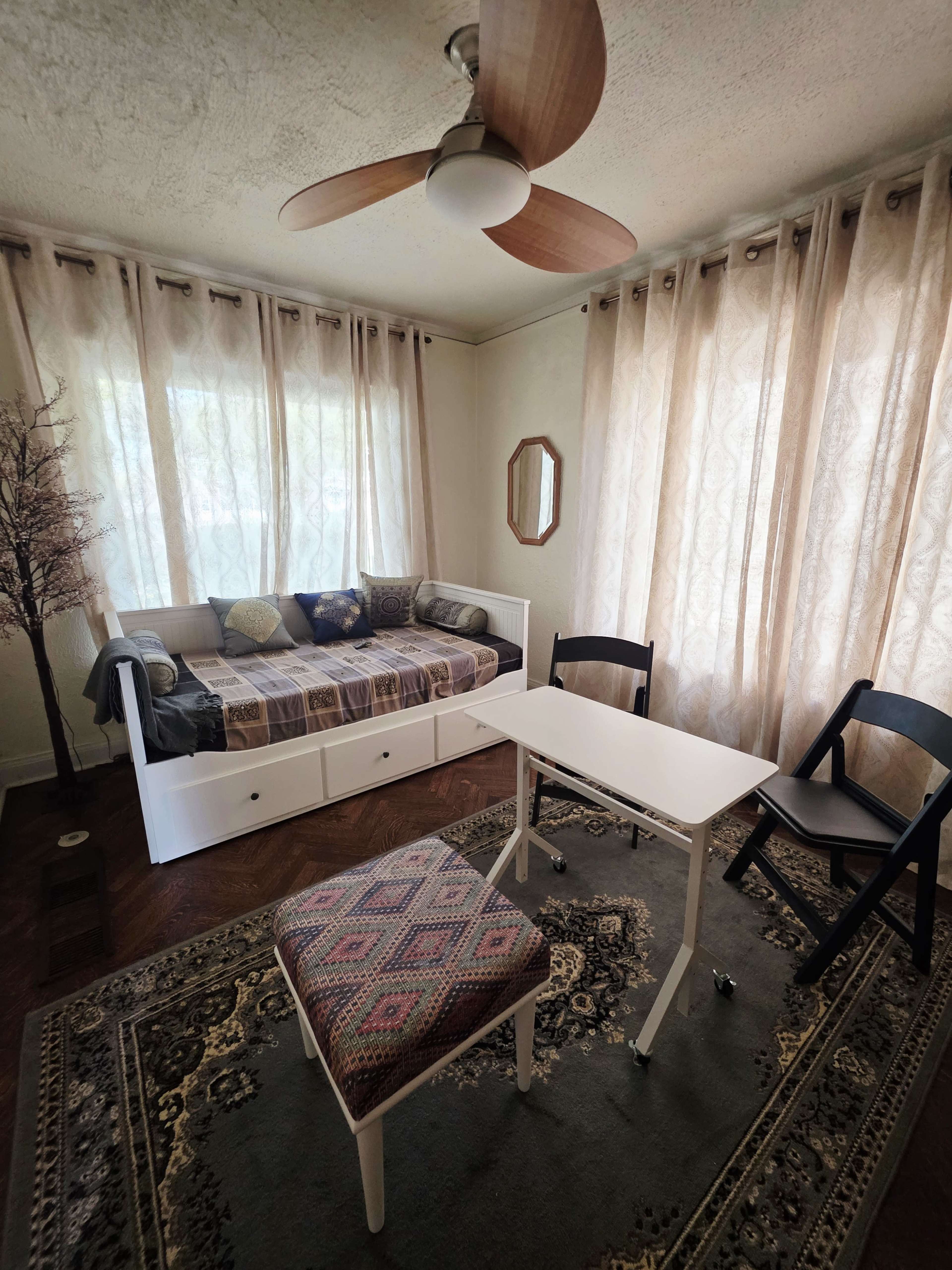 The image shows a well-lit room featuring a daybed with drawers, a small white table, and two chairs on a patterned rug beneath sheer curtains.