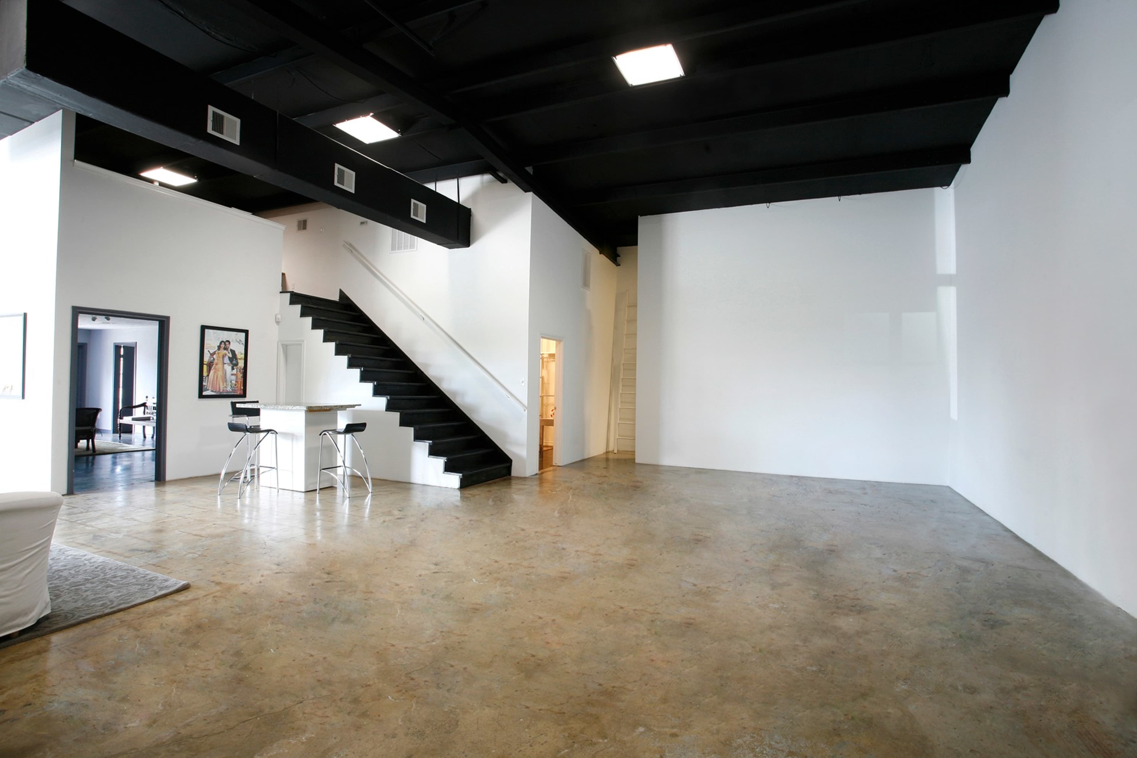 A spacious, minimalist interior featuring a polished concrete floor, a staircase on the left, and a large white wall.