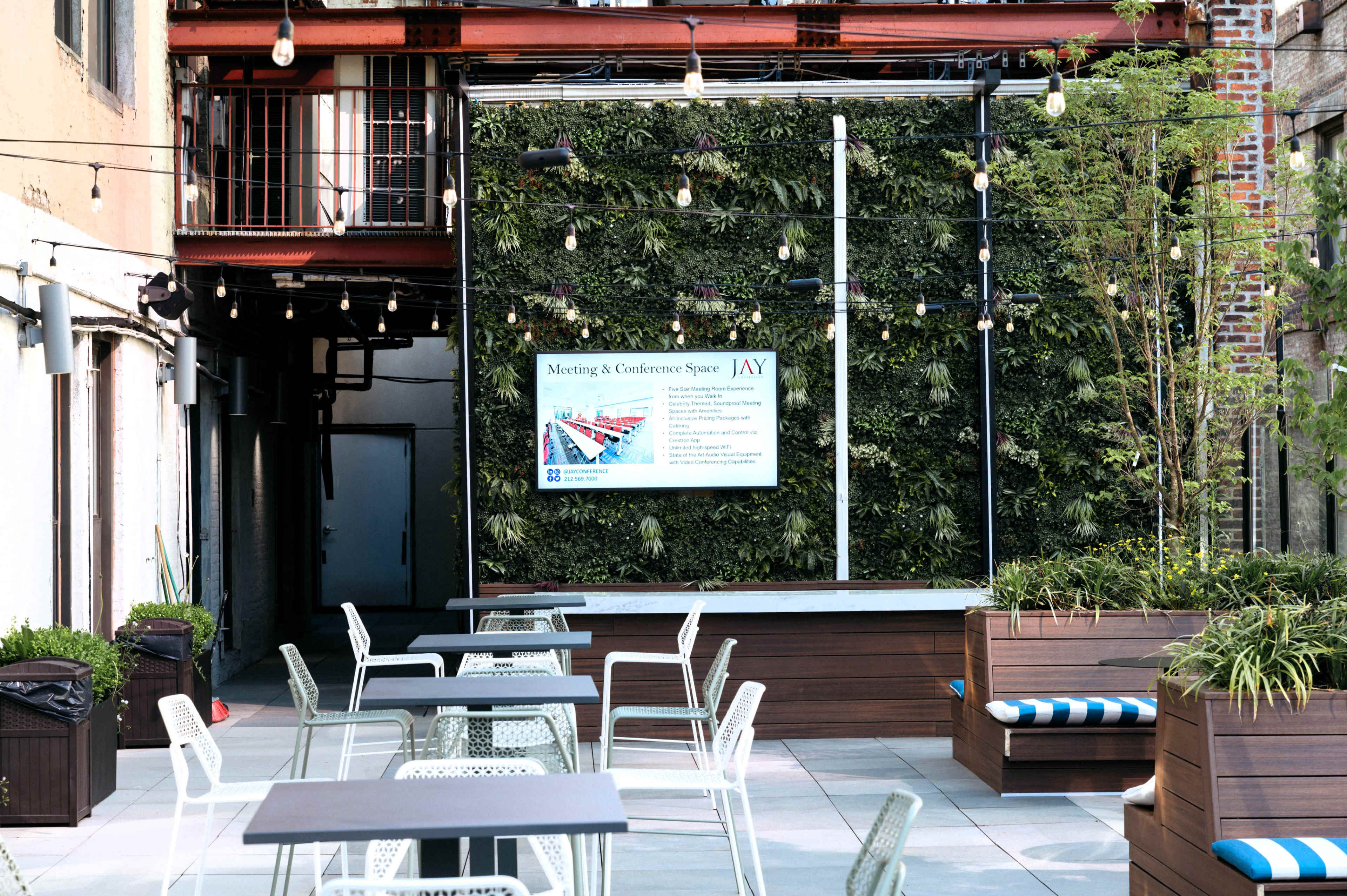 The image shows an outdoor meeting and conference space featuring tables, chairs, and a vertical garden backdrop with string lights overhead.