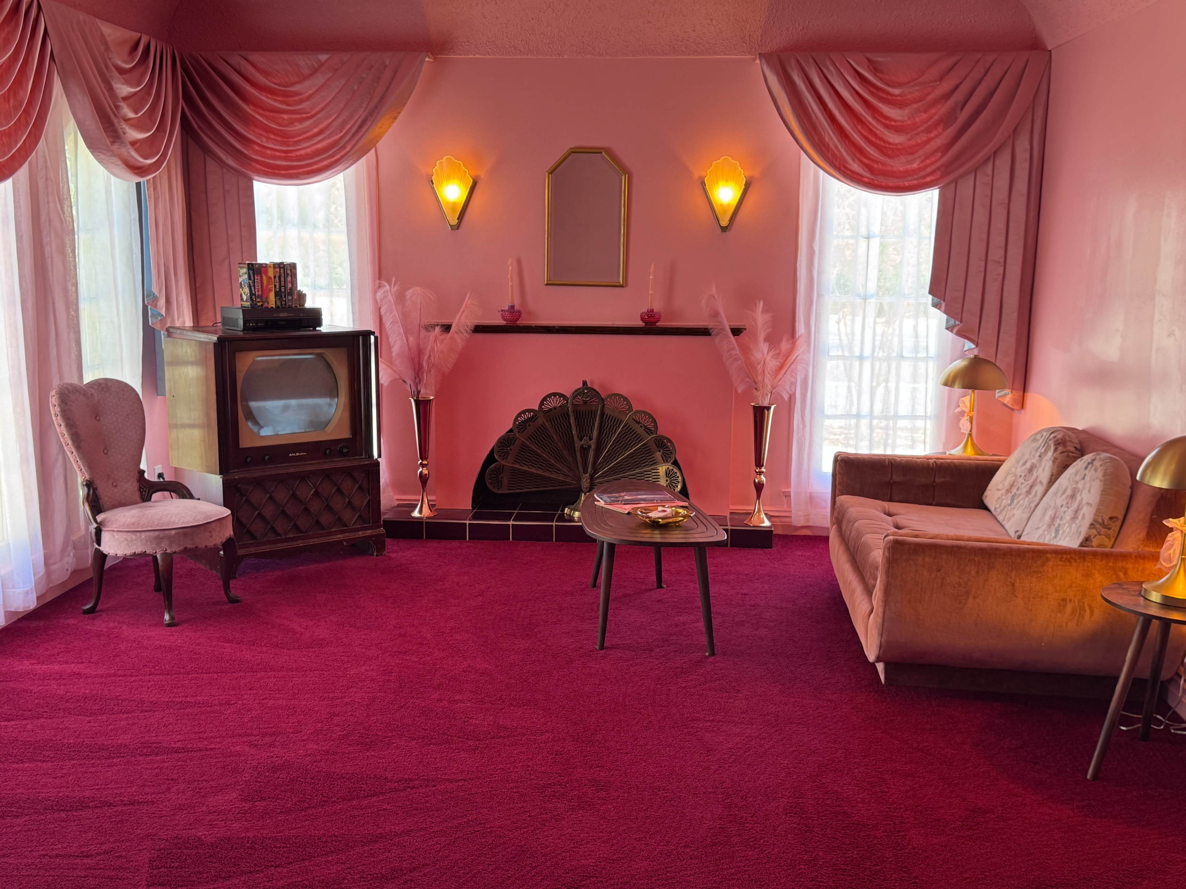 The room features pink walls, a plush carpet, a vintage television, and a combination of seating with a sofa and an armchair, all accented by various decorative elements.