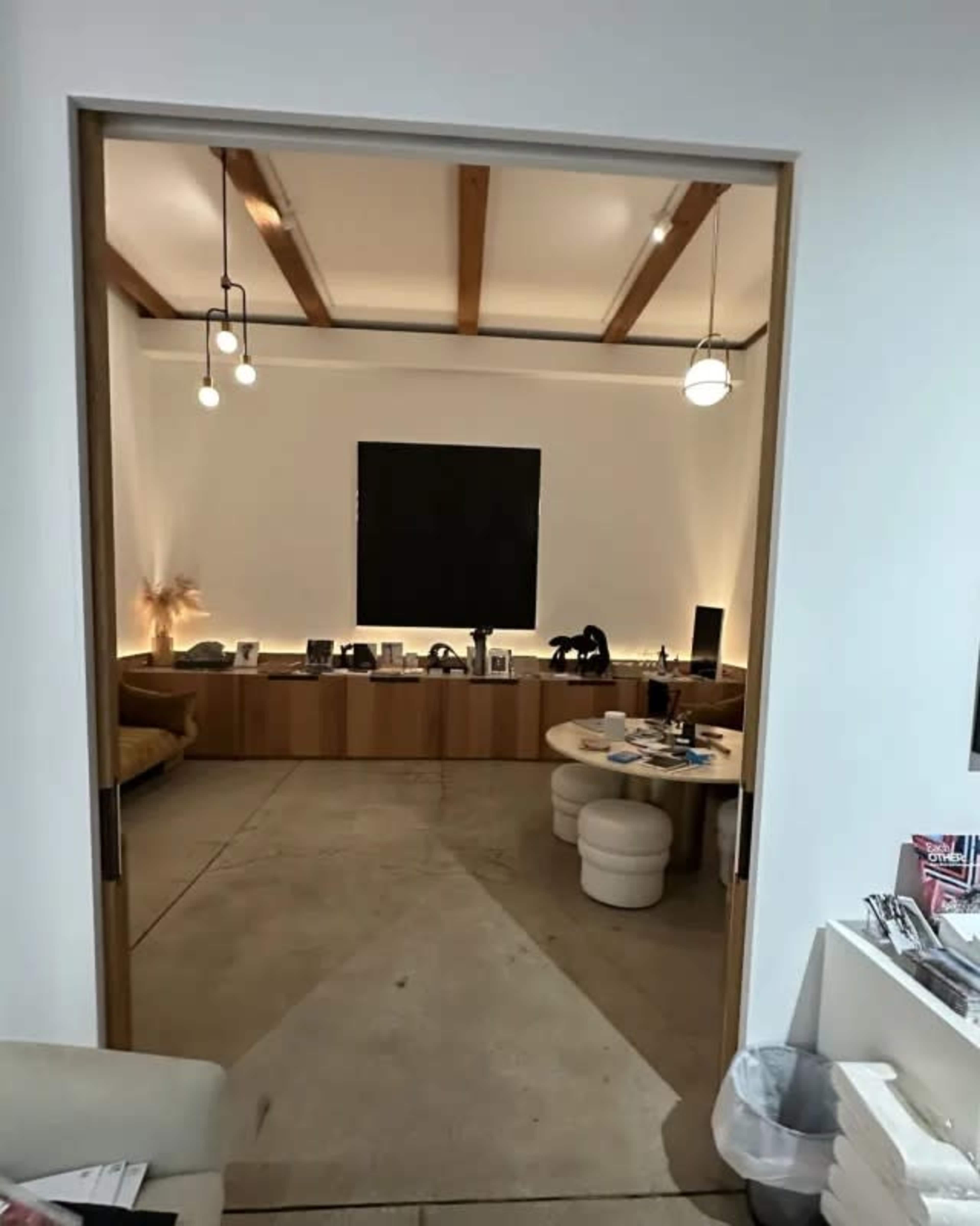 The image shows a room with a minimalist design featuring a sofa, a round table with various items, and a large black wall panel, illuminated by soft lighting.