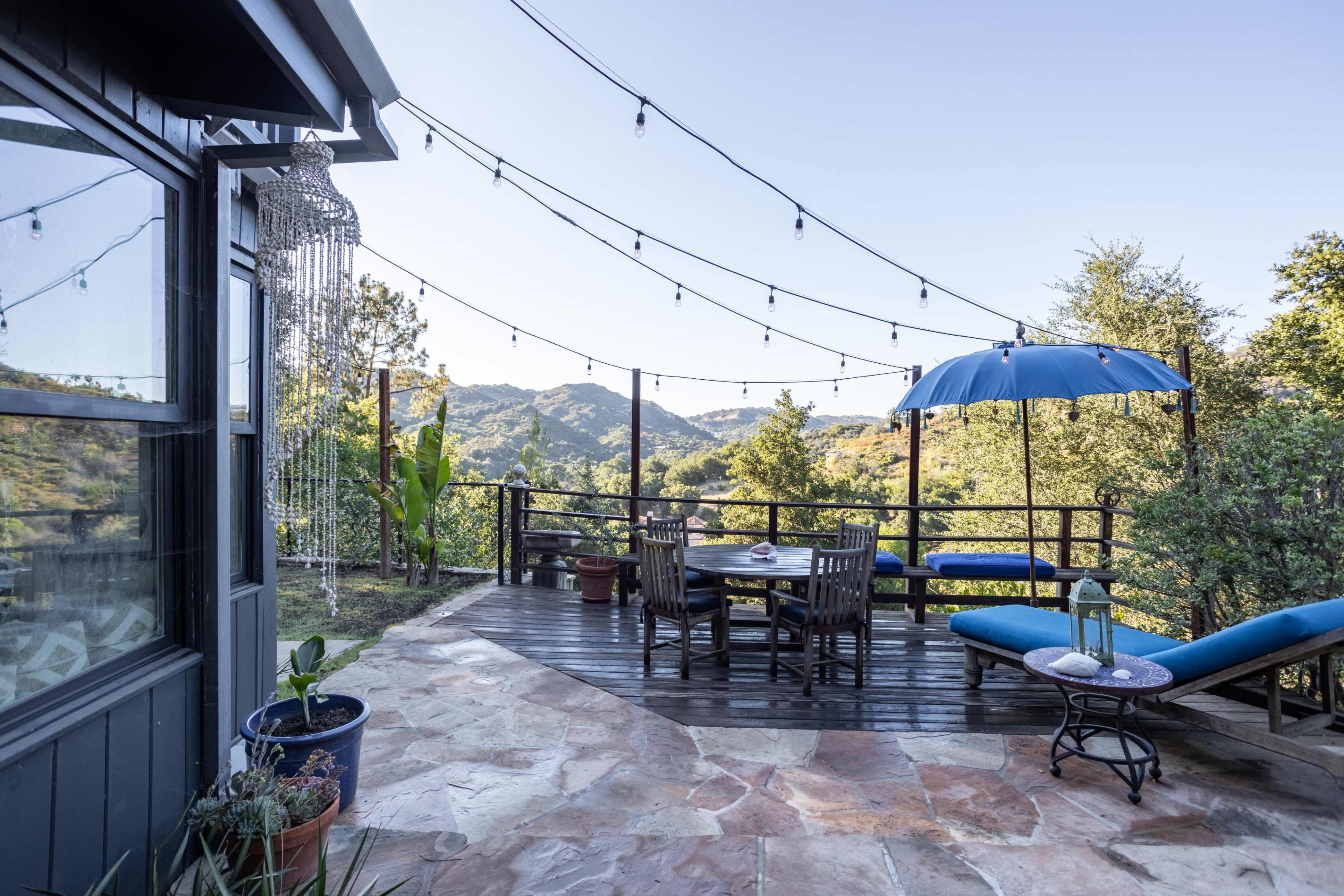 The image shows a patio with a wooden table and chairs, an umbrella, and string lights, overlooking a forested landscape.