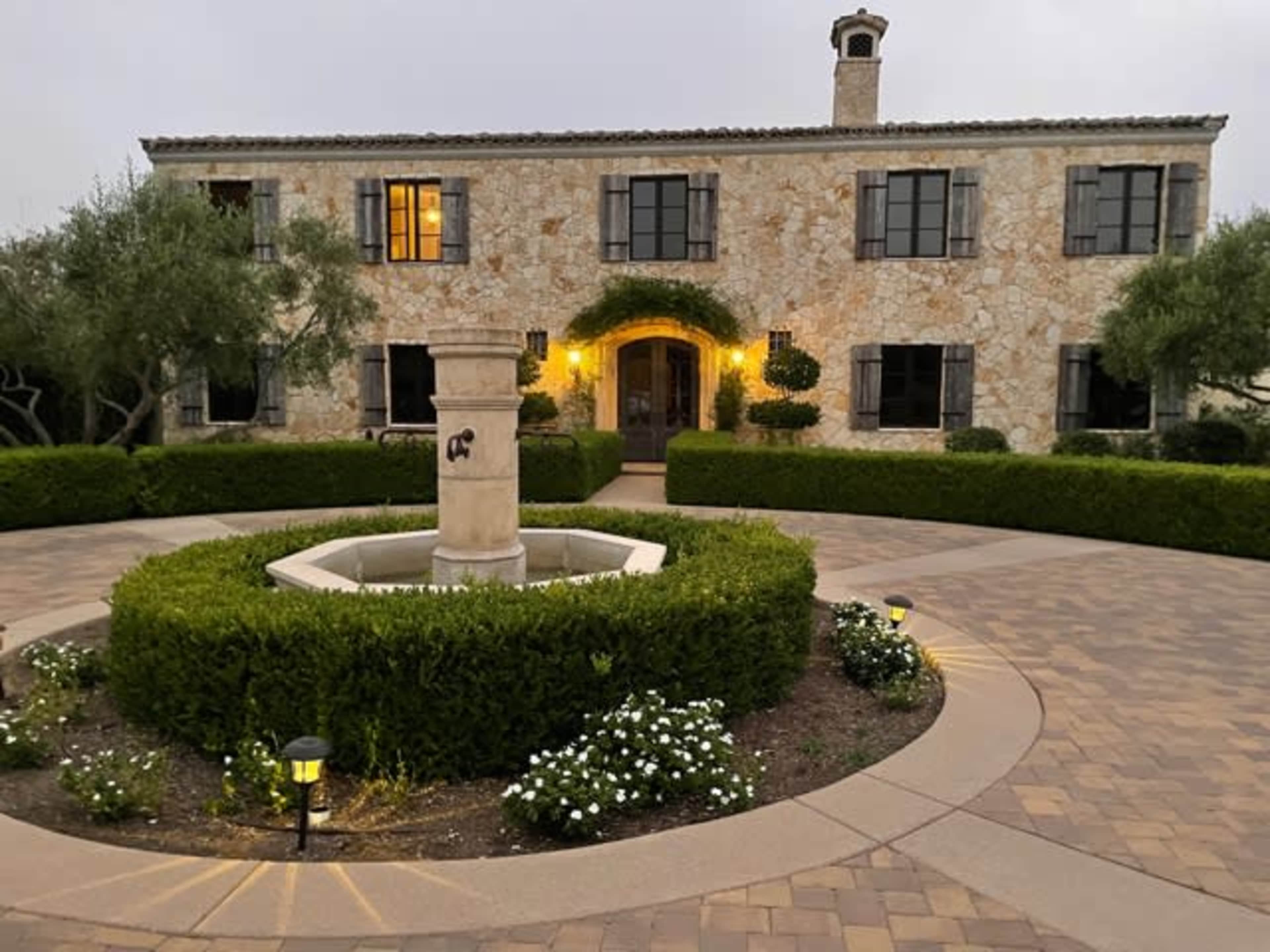 A stone two-story house with dark shutters is surrounded by landscaped gardens and a central fountain in a circular driveway.