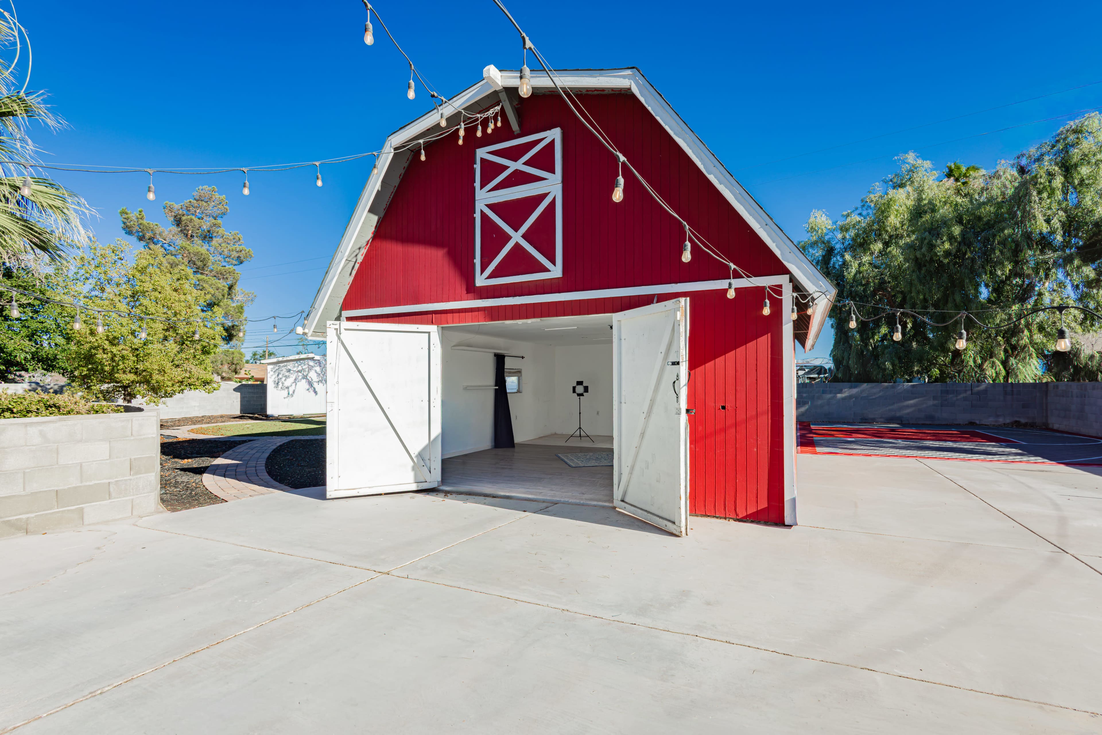 A red barn with open doors is situated on a concrete surface, surrounded by greenery and outdoor string lights.