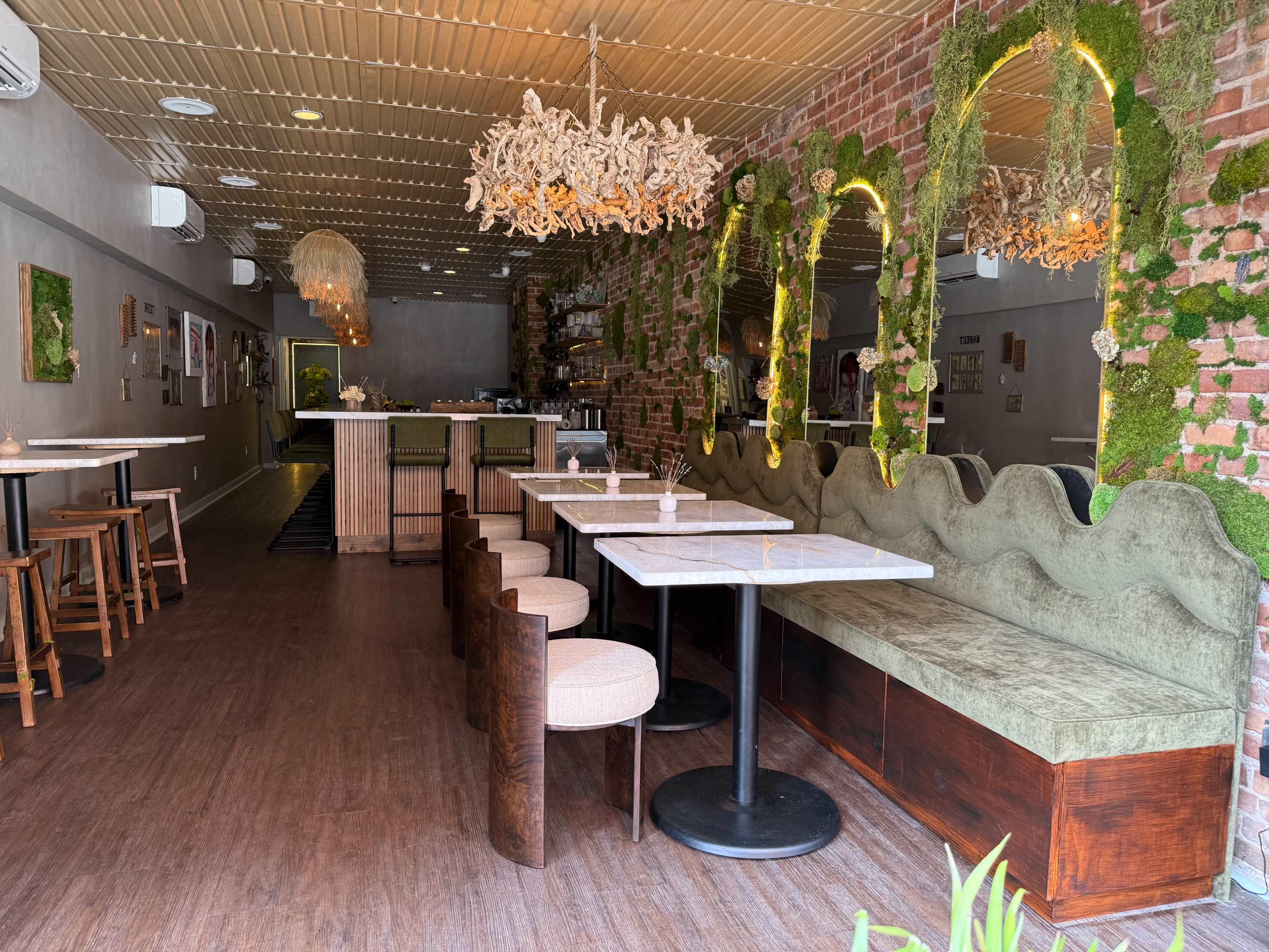 The image shows a modern restaurant interior featuring wooden tables and chairs, a large upholstered bench along one wall, and greenery decorations hanging from the ceiling and walls.