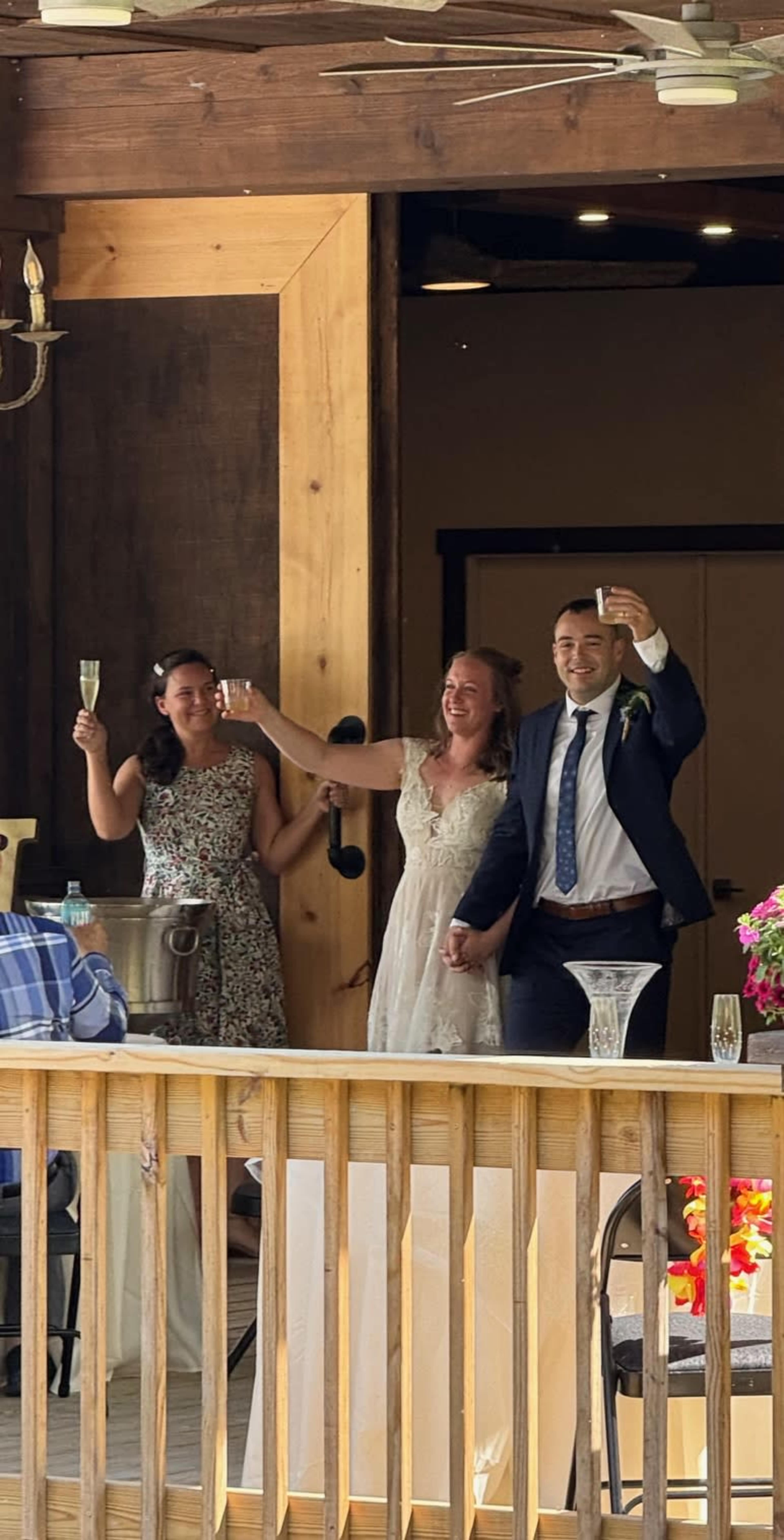 A bride and groom hold hands and raise champagne flutes in celebration alongside a woman in a floral dress at a reception venue.