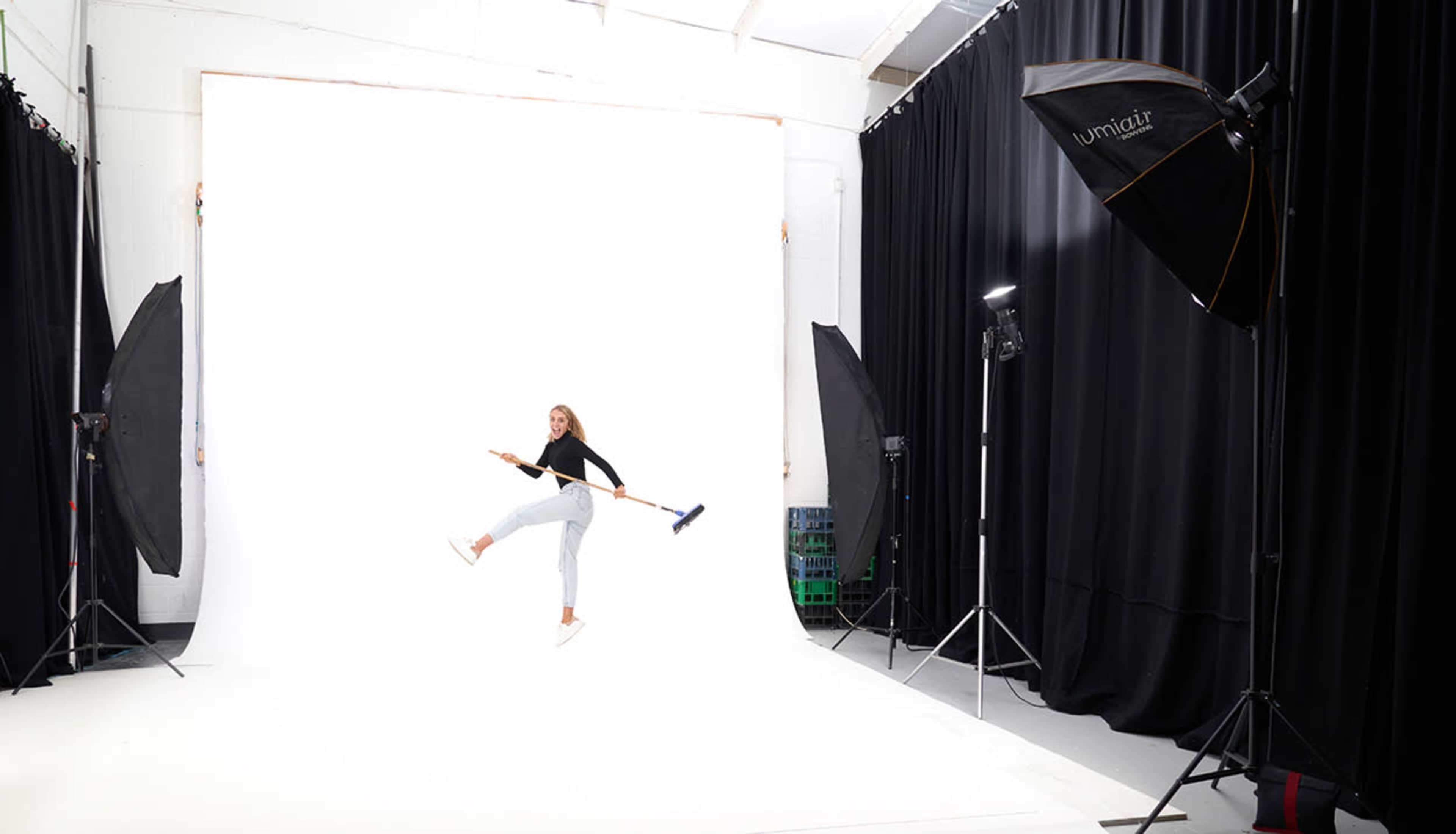 A person is playfully holding a broom while jumping in a photography studio with a white backdrop and lighting equipment.