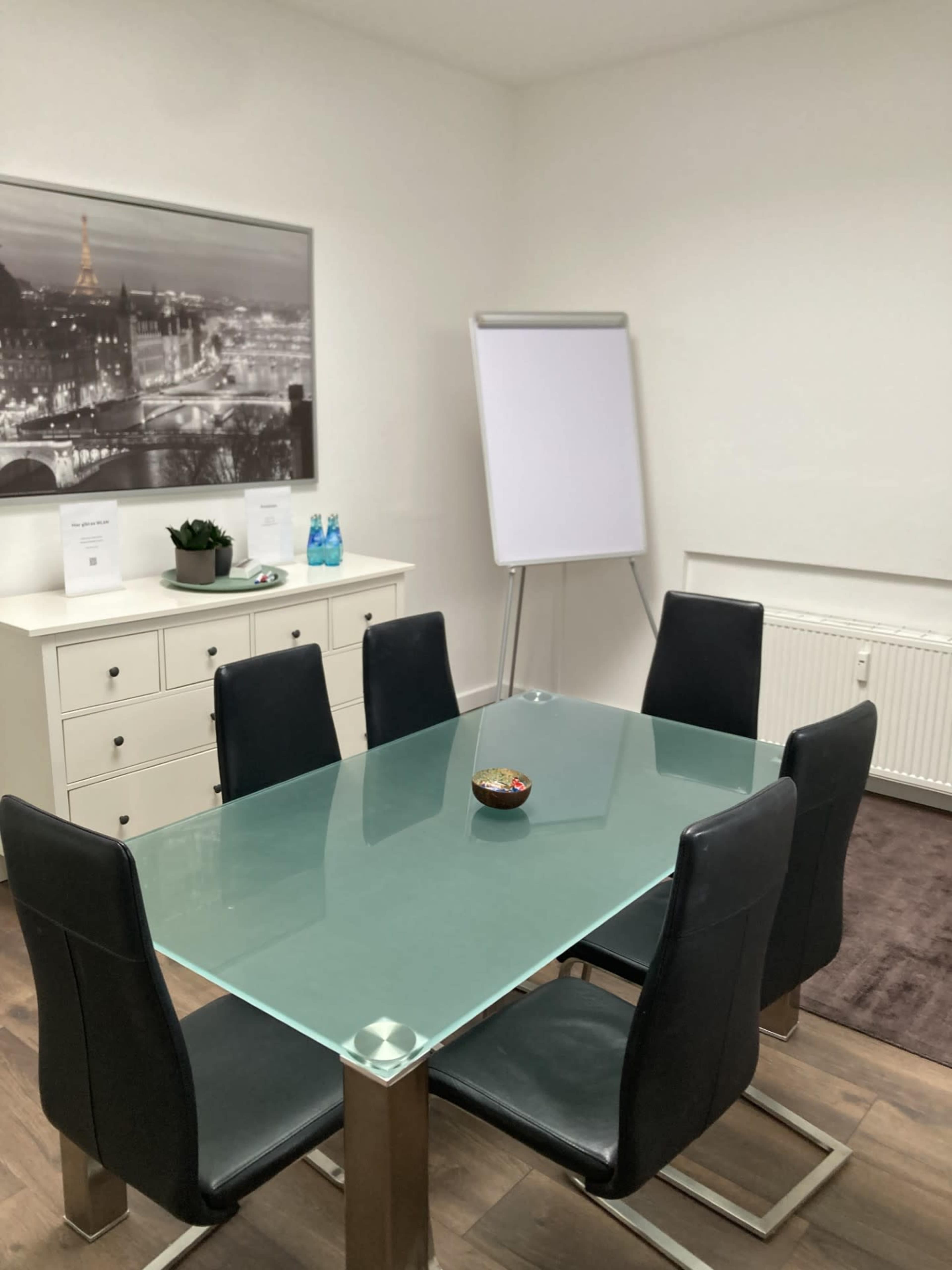 The image shows a small conference room with a glass-top table, six black chairs, and a whiteboard, along with a plant and water bottles on a cabinet.