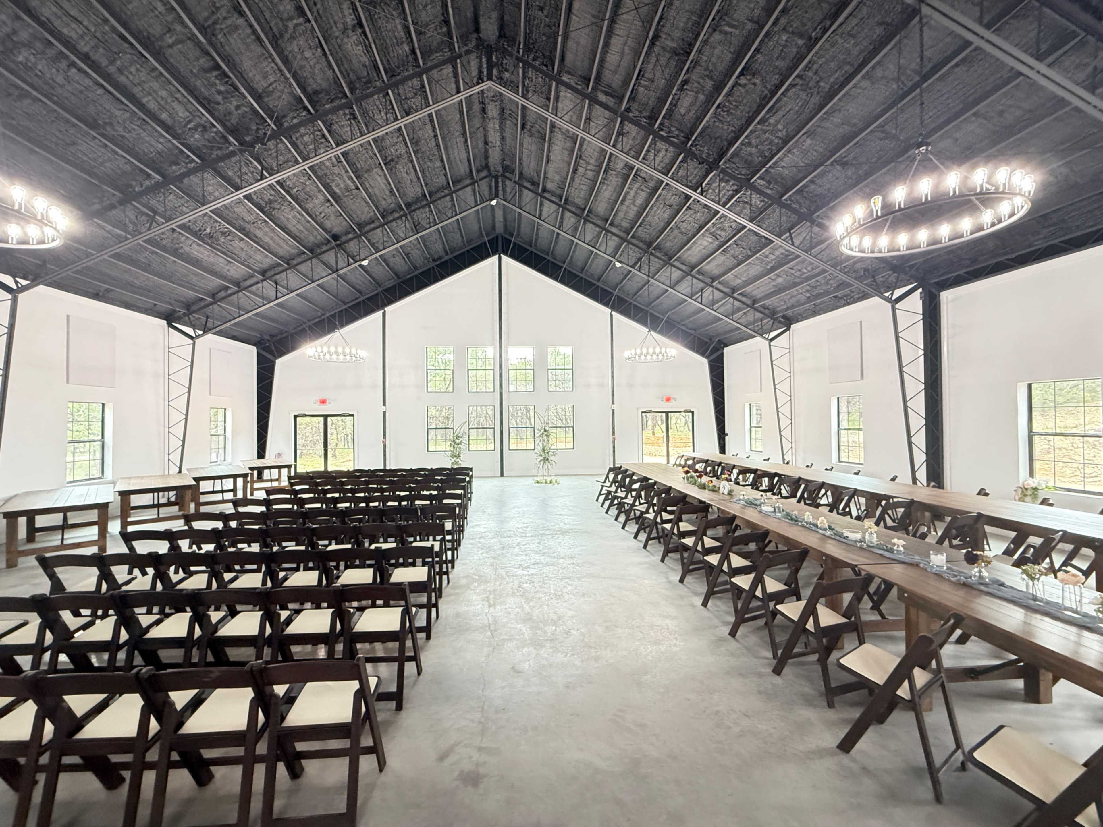 The image shows a spacious, modern event venue with rows of wooden chairs facing a long dining table under a high ceiling with chandeliers.