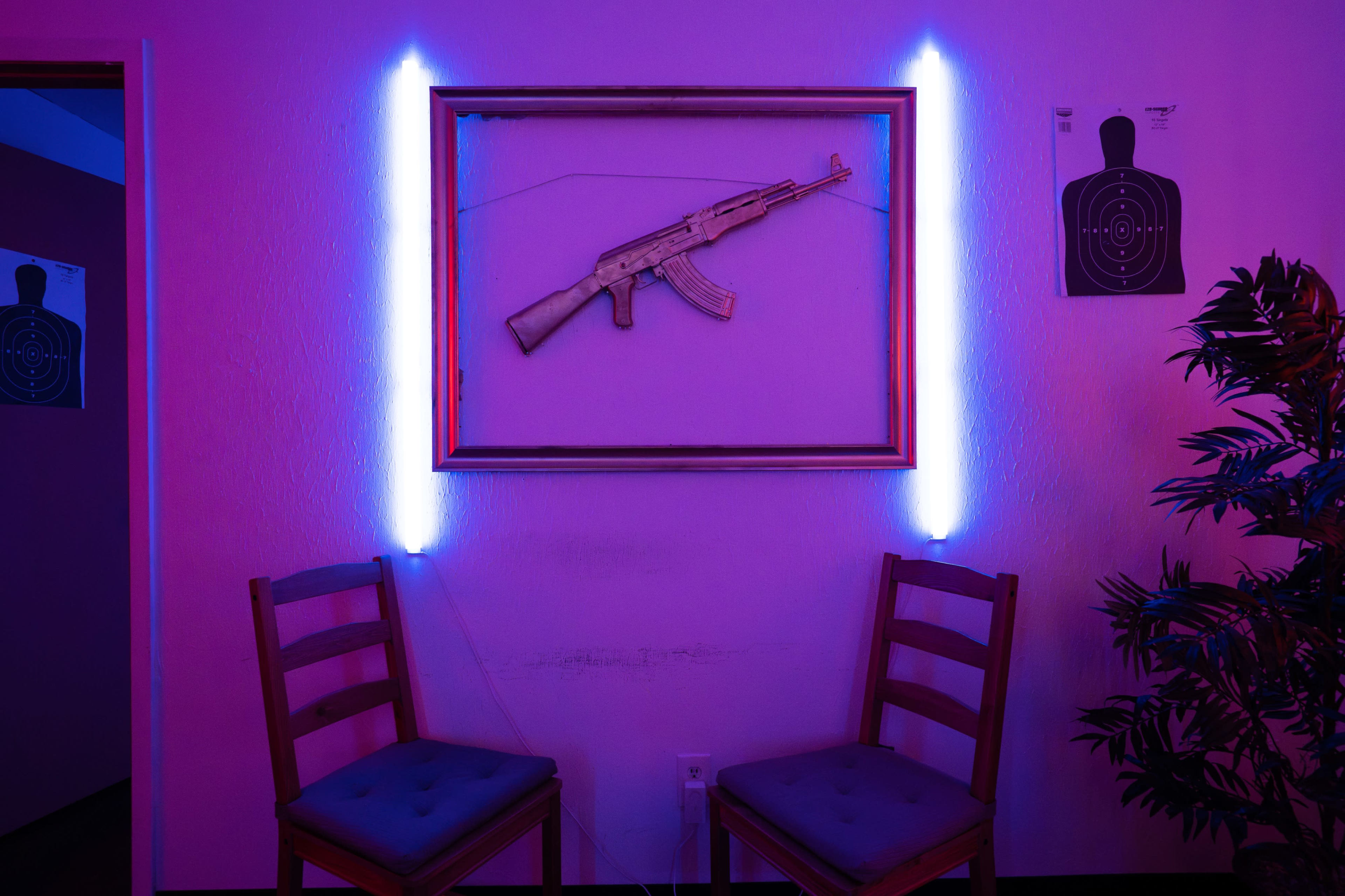 A framed assault rifle is displayed on a wall between two illuminated vertical light strips, with two chairs positioned in front and target practice sheets on the adjacent wall.