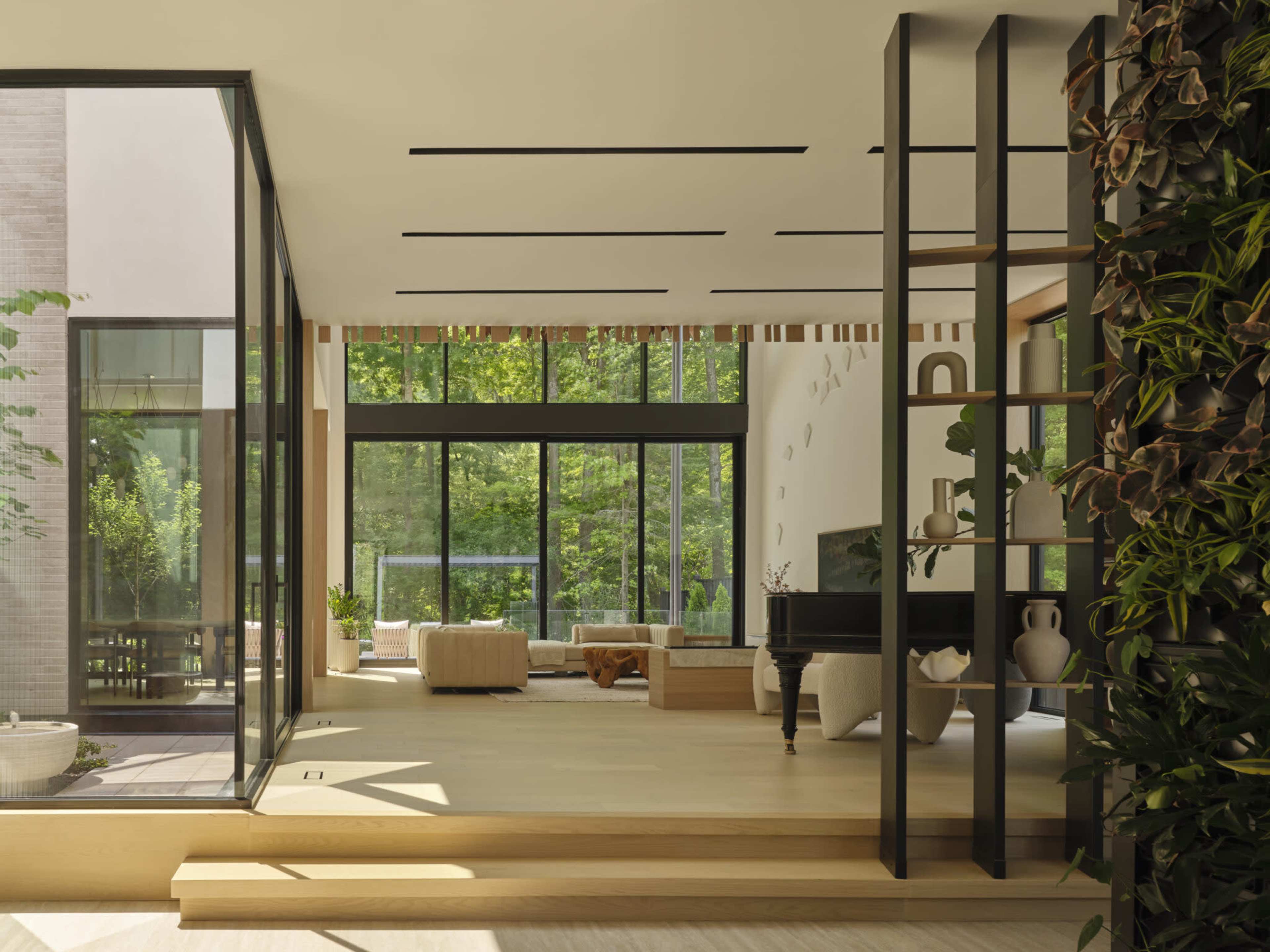 A bright, modern living space features large glass windows, a piano, and a greenery wall divider.
