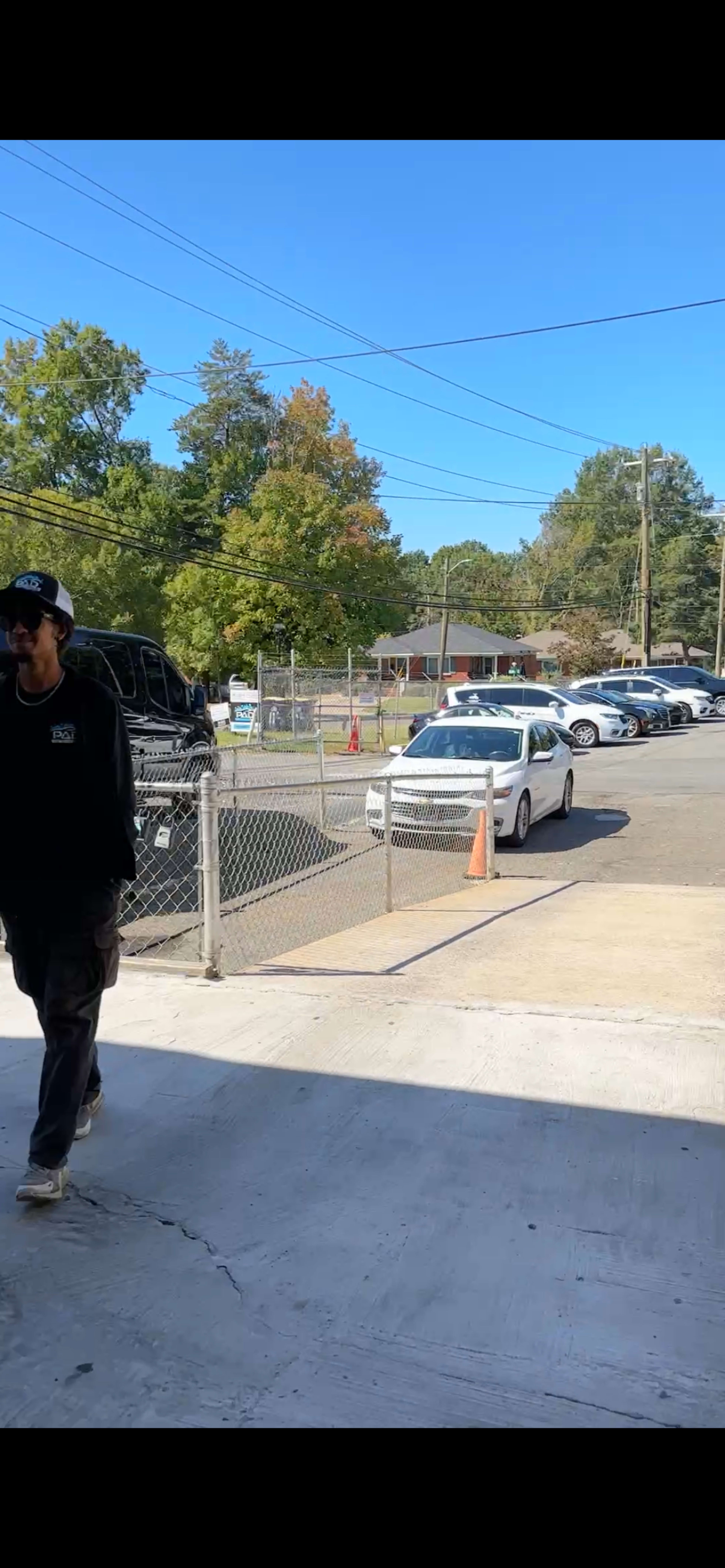 A man wearing a dark sweatshirt walks past a parking lot filled with parked vehicles under a clear blue sky.